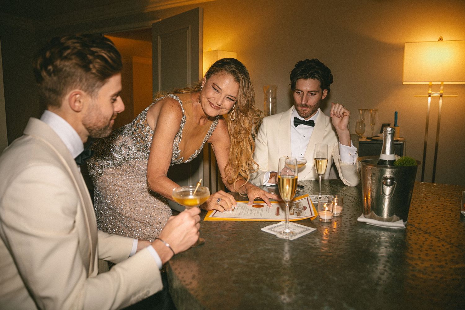Wedding guests in formal attire socialize at a bar with champagne during a reception.