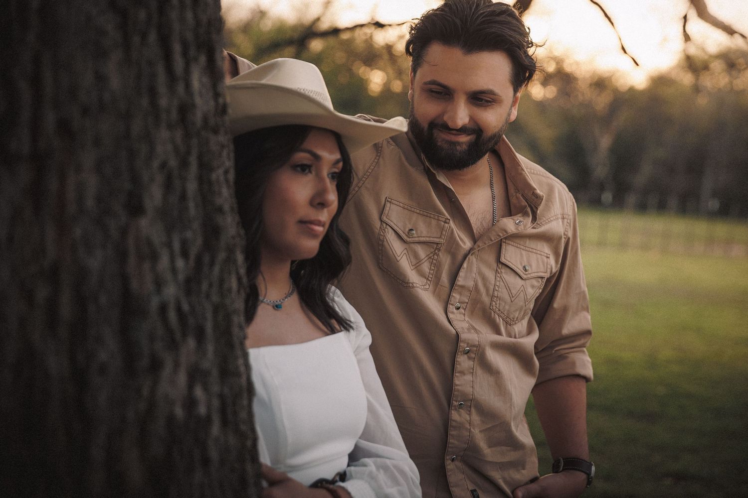 Couple leaning against a tree at sunset, woman in white top and cowboy hat with man in tan shirt.