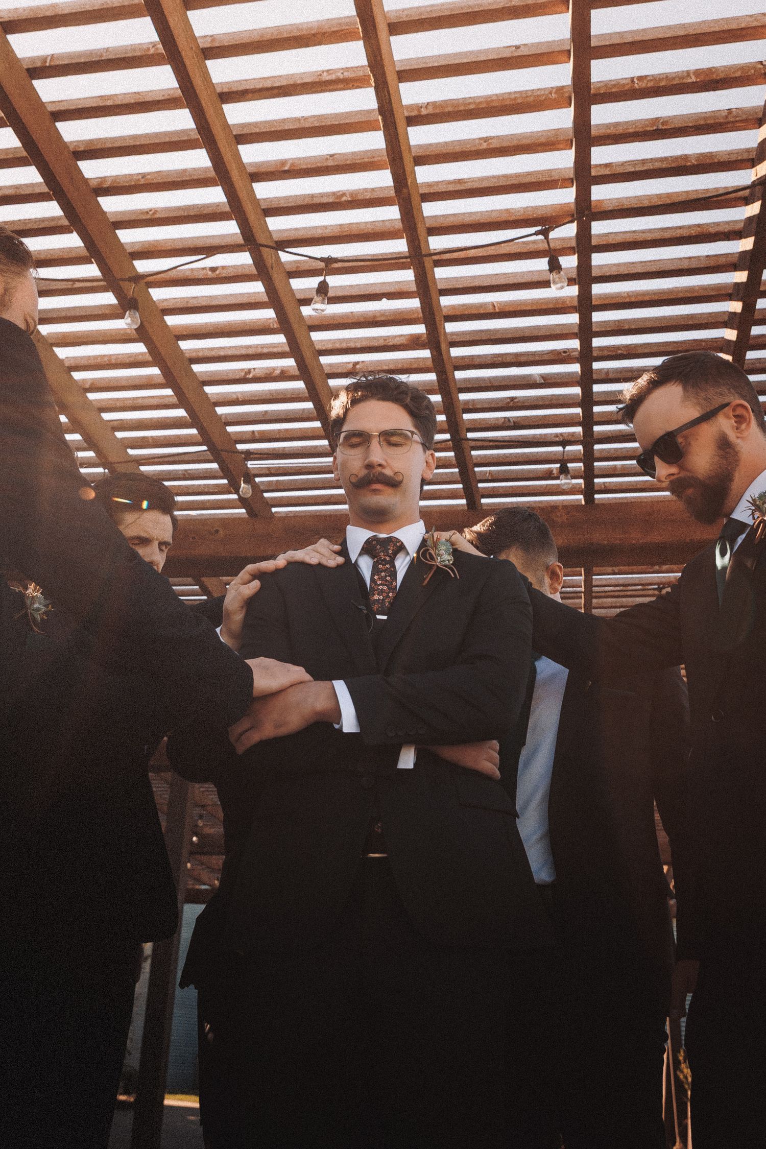 Groom with mustache stands arms crossed surrounded by groomsmen under wooden pergola.