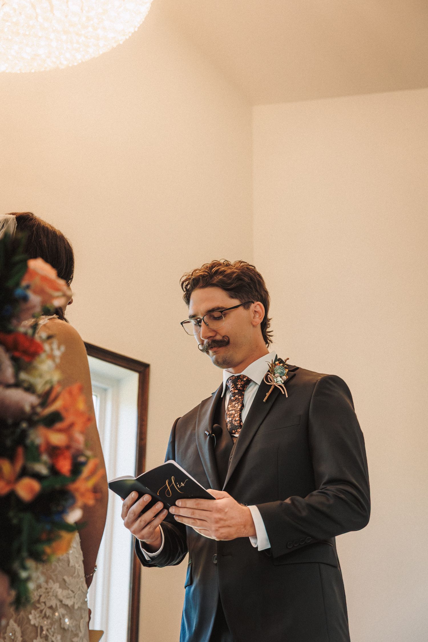 Groom with mustache and glasses reads vows from a booklet while wearing a floral tie and boutonniere.