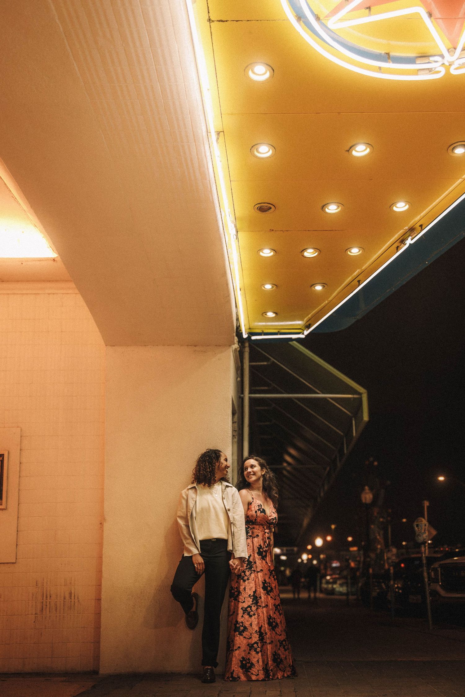 Engaged couple leans against wall under bright theater marquee lights at night, woman in floral maxi dress.