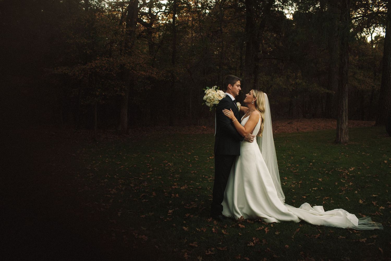 Bride and groom share intimate moment surrounded by tall autumn trees in moody evening light.