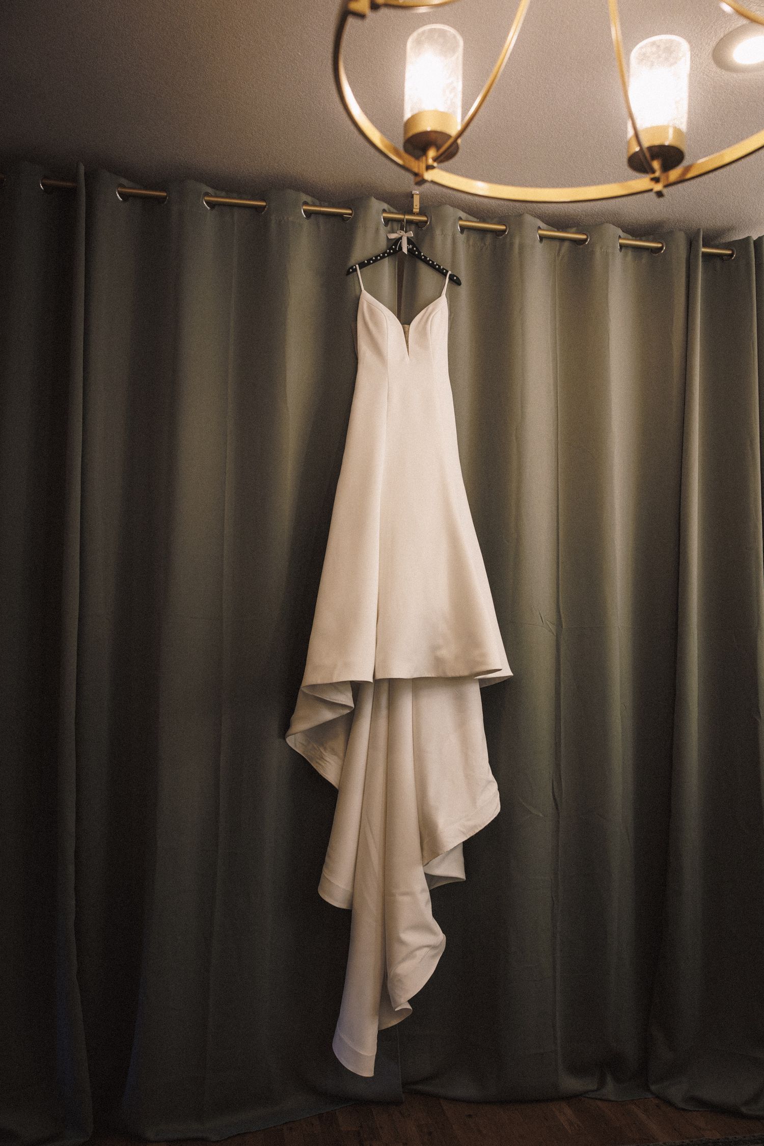 Elegant white satin wedding dress with asymmetrical hem hanging from gold chandelier against gray curtains.