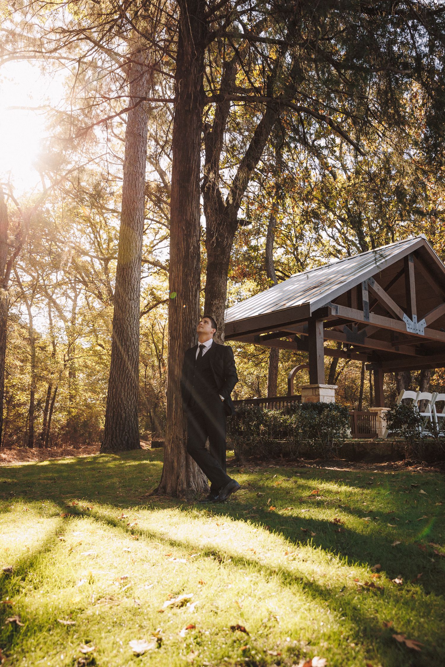 Groom in dark suit leaning against tall tree in sunlit outdoor garden with rustic pavilion backdrop.