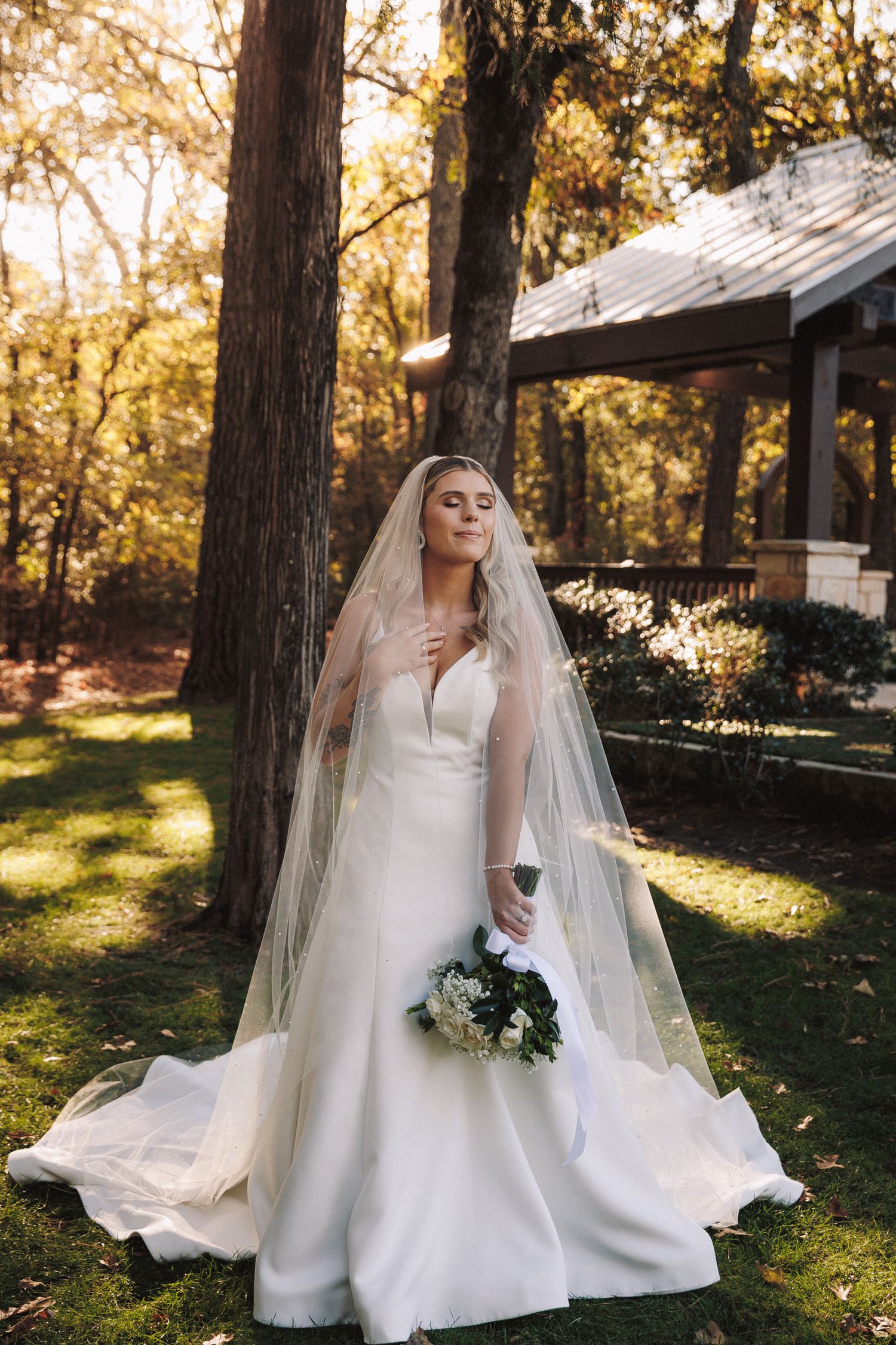 Bride in white gown and veil holding bouquet standing by trees near pavilion.