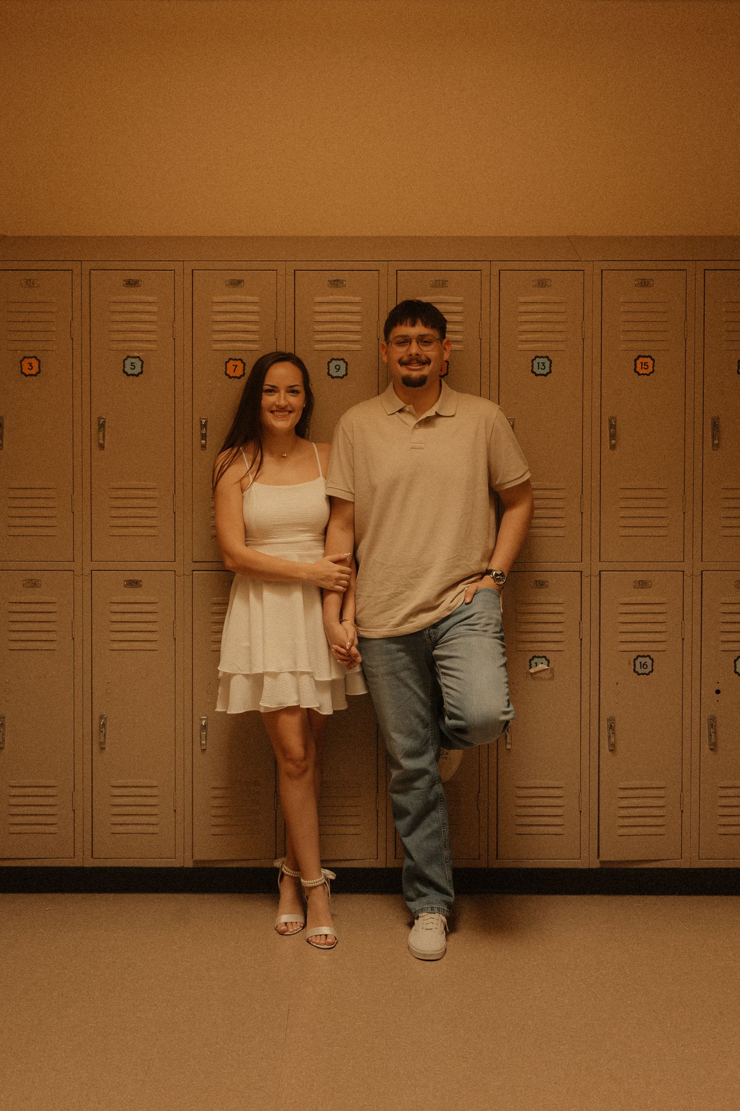 Smiling couple holding hands and posing in front of beige school lockers in a warmly lit hallway.