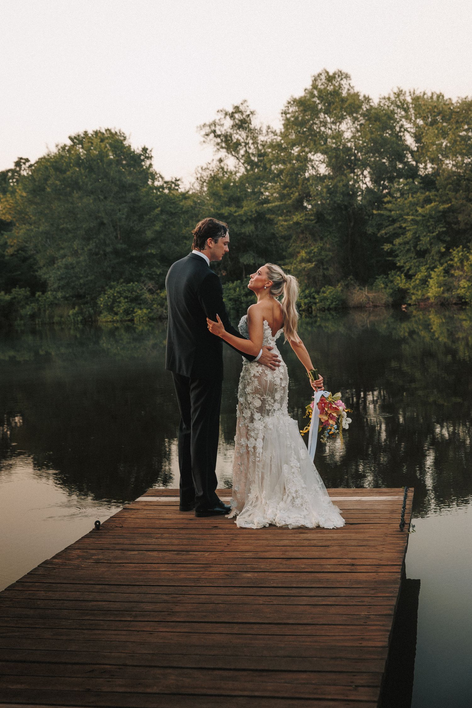 A wedding couple stands on a wooden dock overlooking a tranquil lake during golden hour.