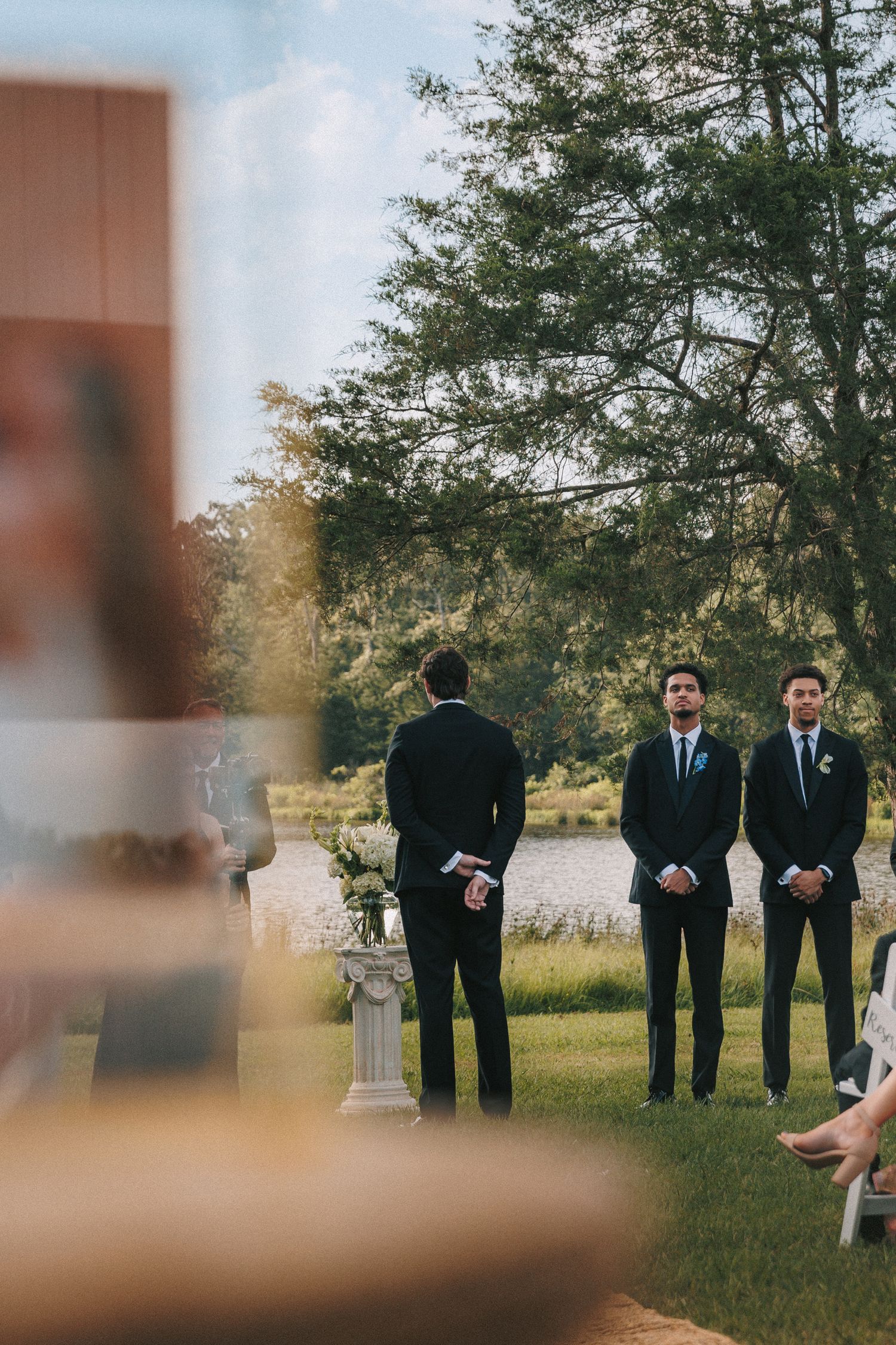 Groomsmen in dark suits stand in formation during outdoor ceremony.