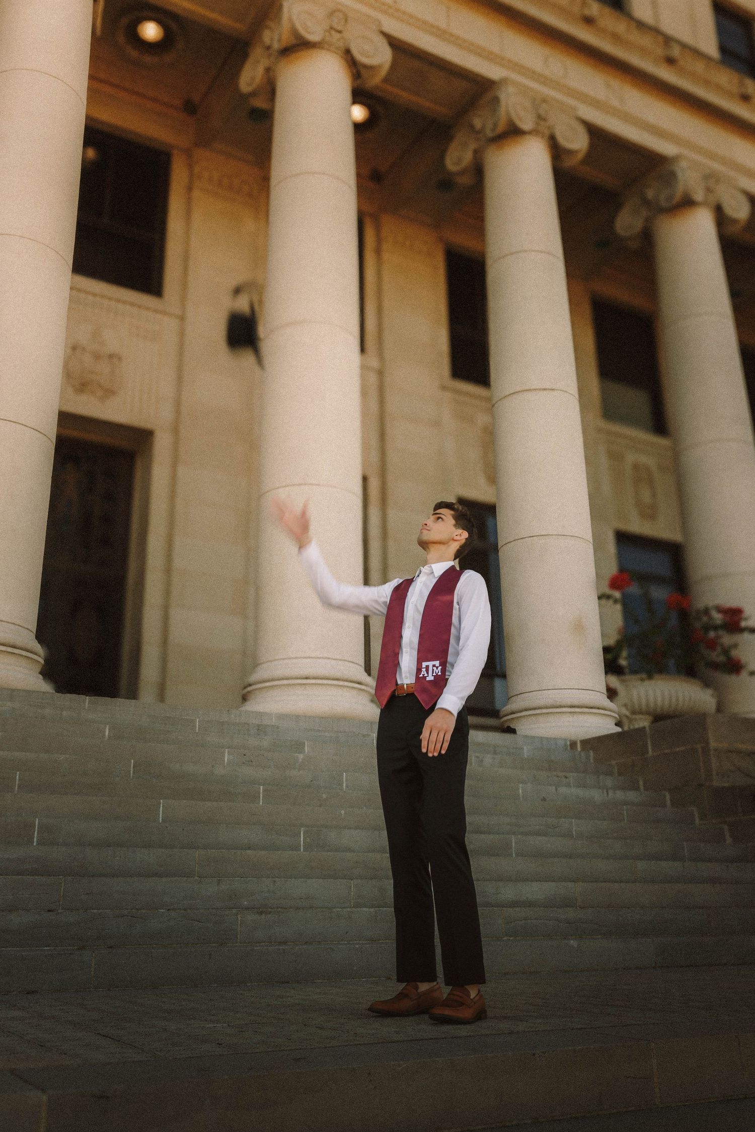 A person in formal attire poses dramatically on stone steps against a backdrop of elegant classical columns.