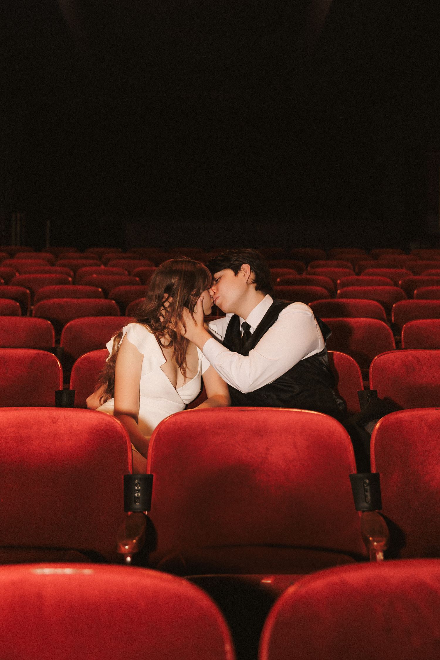 A silhouetted couple shares an intimate kiss in a dark, empty movie theater with rows of red velvet seats.