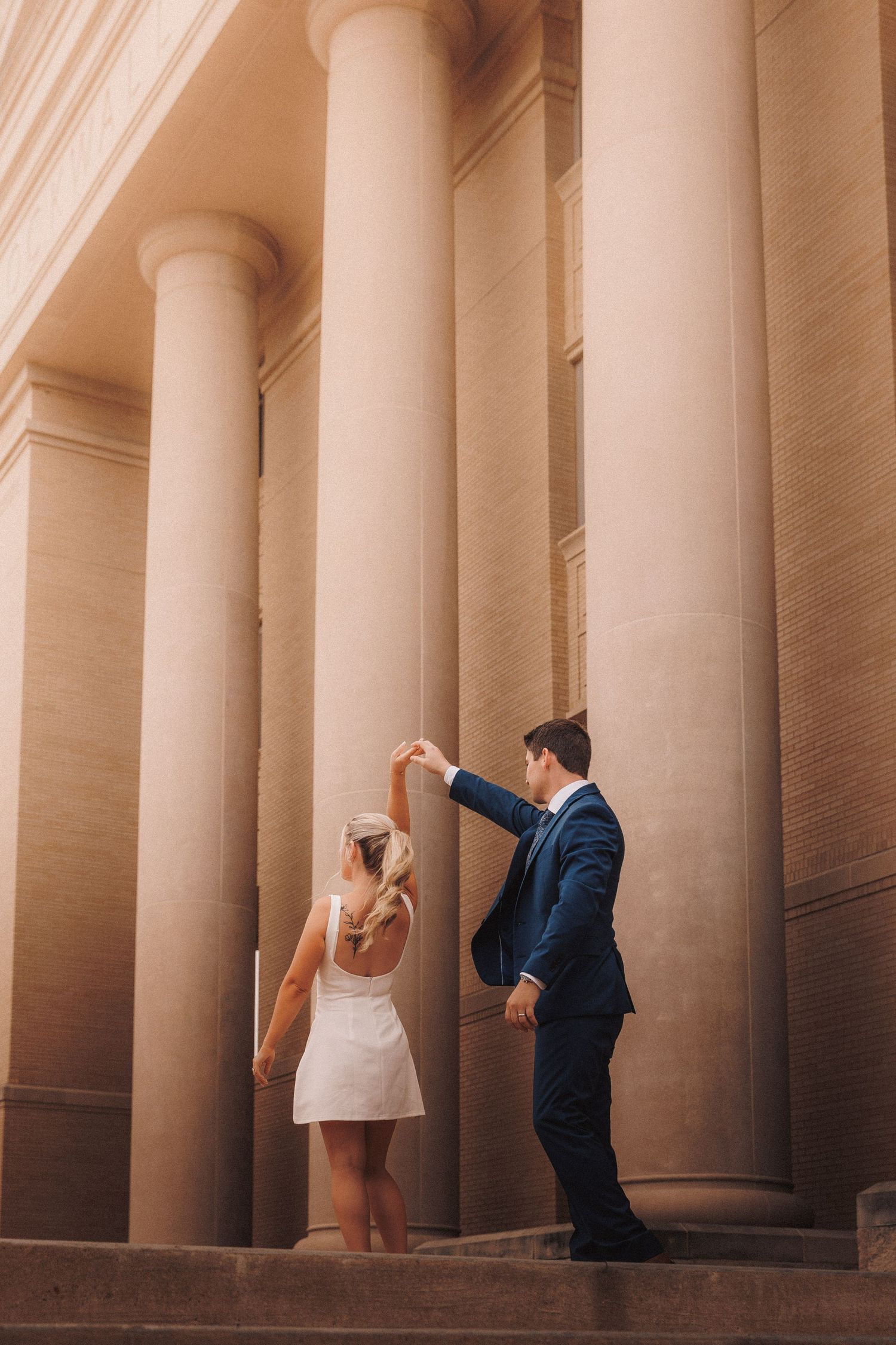 Couple in formal attire pose creatively between grand architectural columns in a romantic photoshoot setting.
