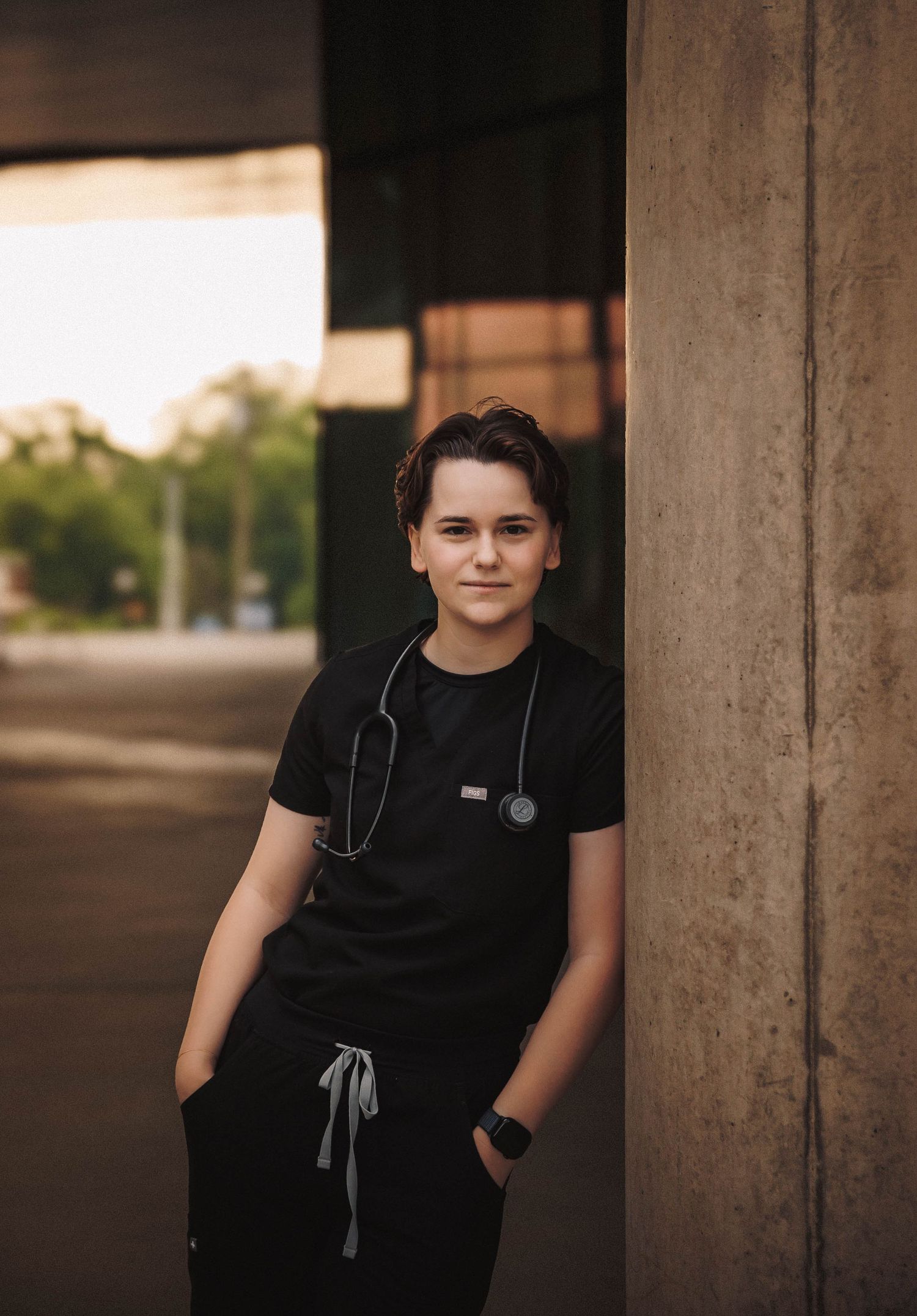 Medical professional in black scrubs and stethoscope standing in front of columns during outdoor portrait session.