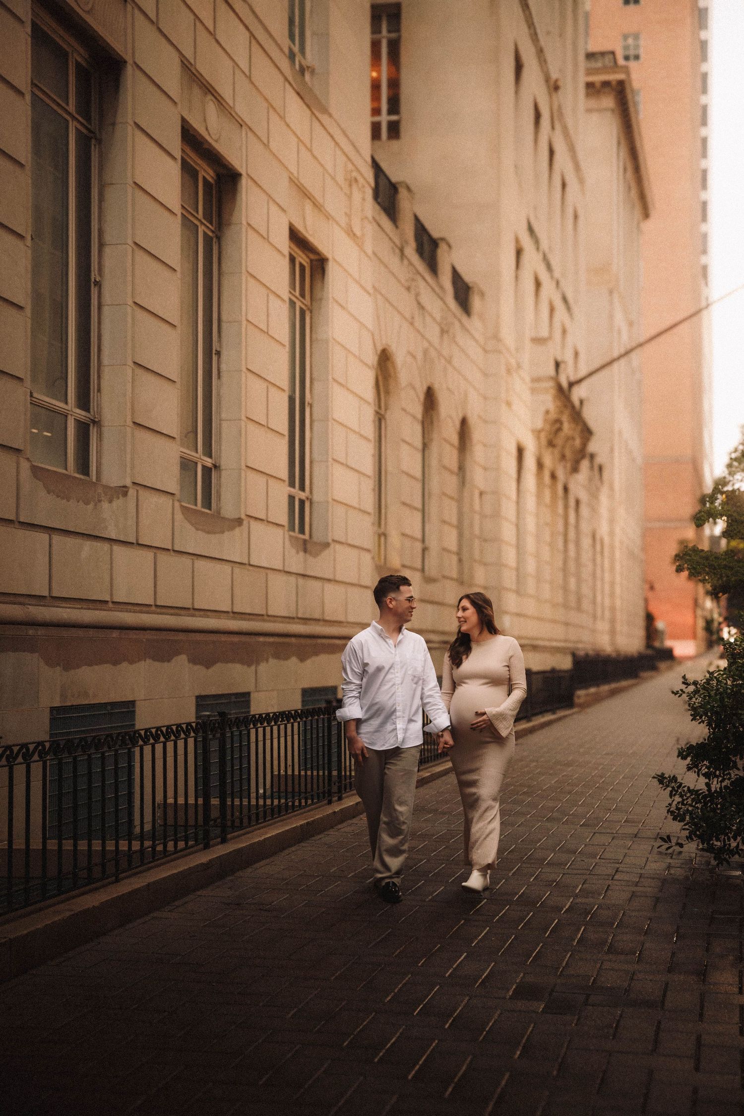 A couple walks along a sloped urban pathway bordered by beige stone buildings in warm evening light.