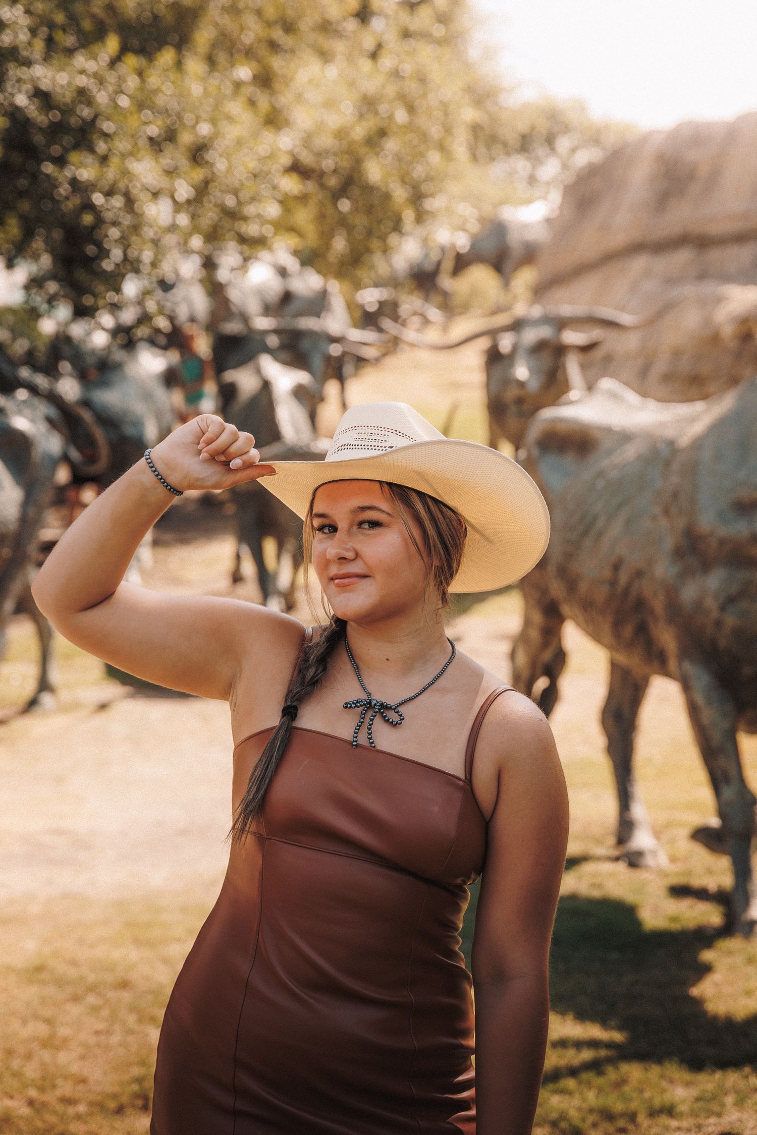 Woman in a brown dress and sun hat smiles while shielding her eyes on a sunny summer day outdoors.