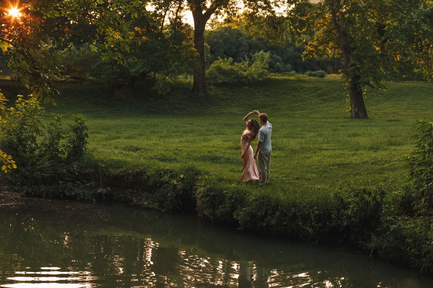 Two people embrace at sunset in a lush garden setting beside a tranquil pond with golden sunlight filtering through trees.