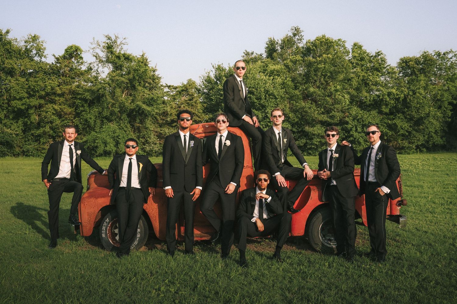 Groomsmen in dark suits pose together on green lawn for formal wedding photos.