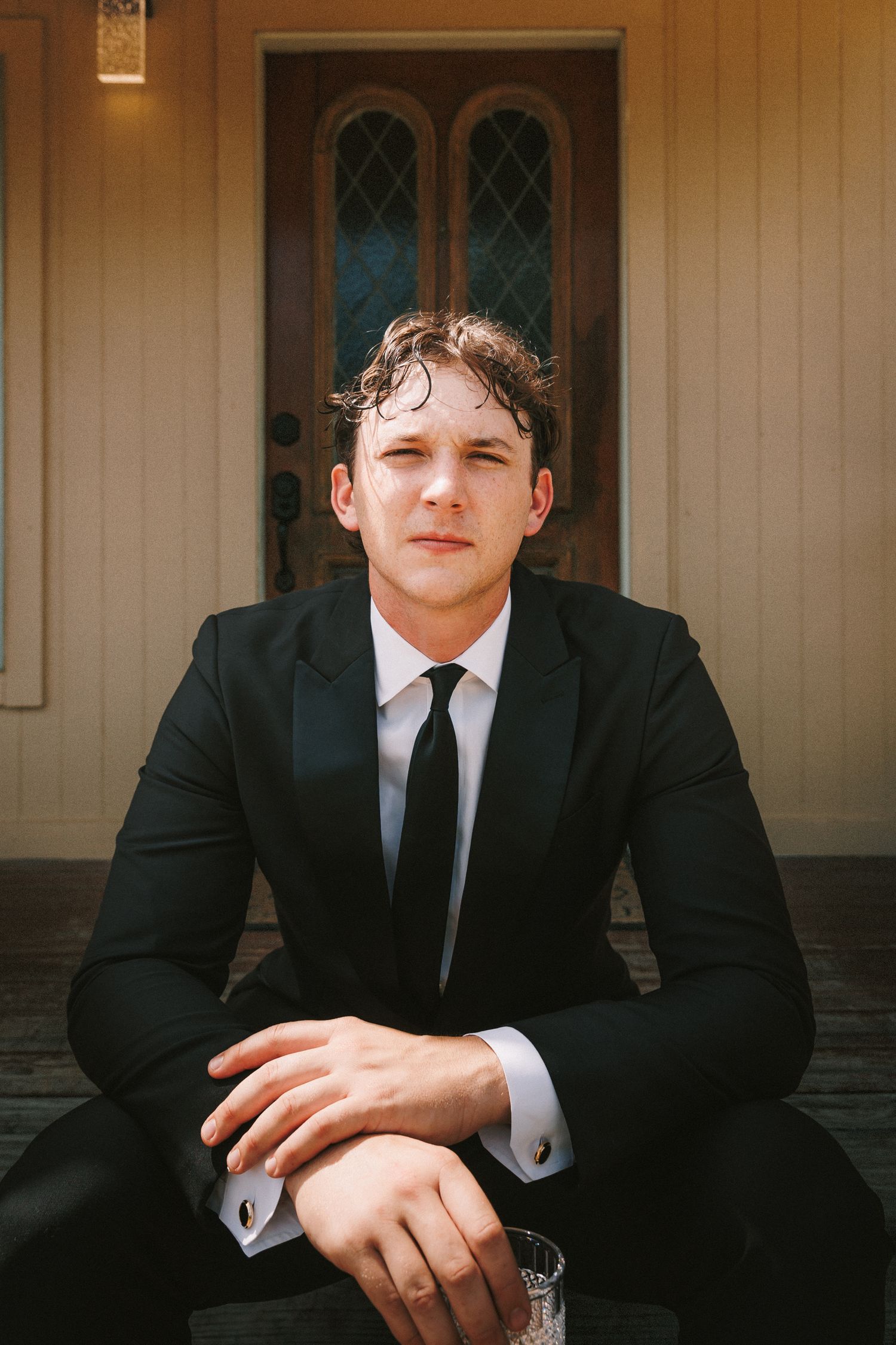 A person seated wearing a formal black suit and tie against a beige building exterior.