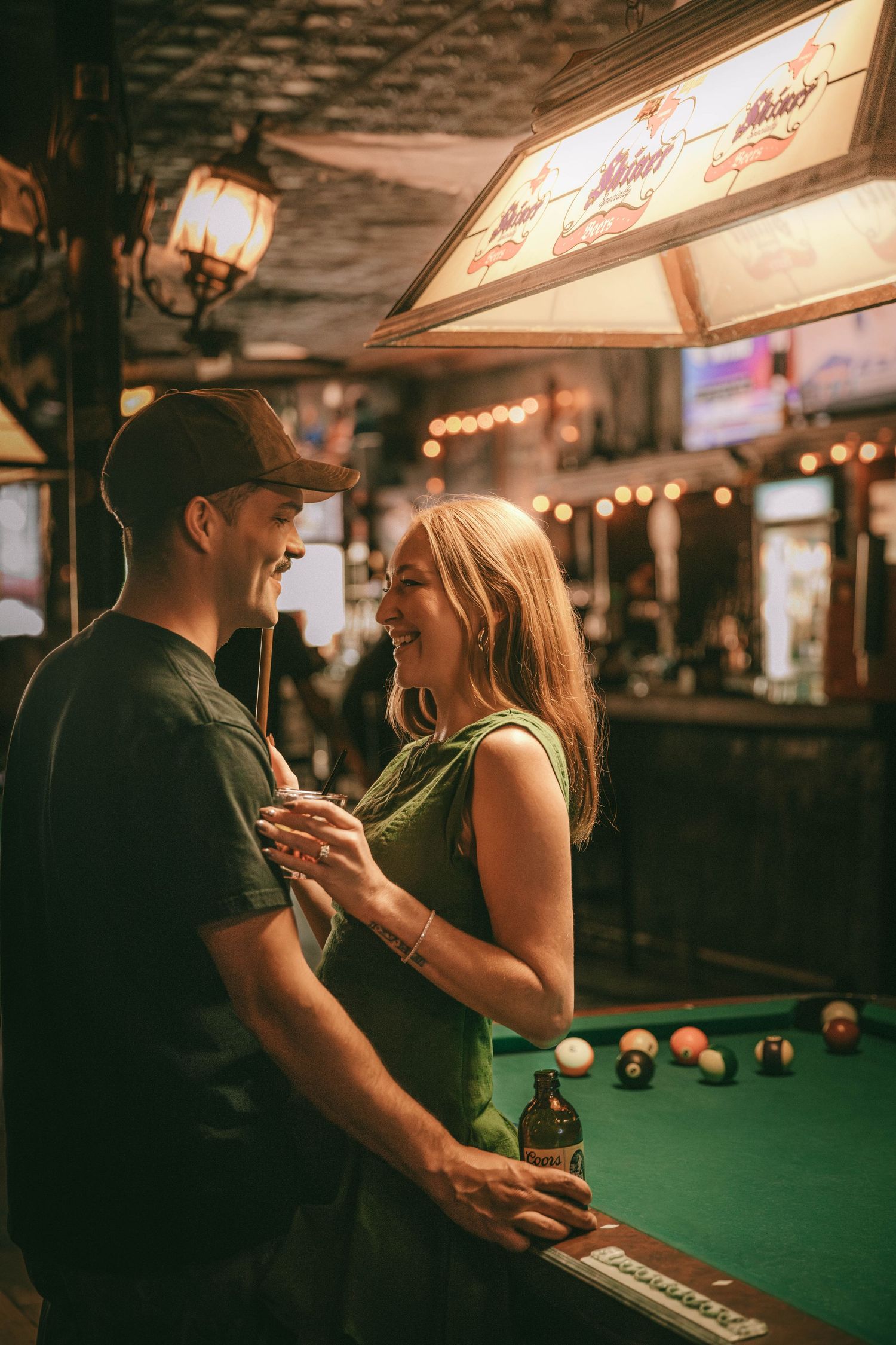 A couple shares an intimate moment while playing pool at a dimly lit bar with green felt tables and neon signs.
