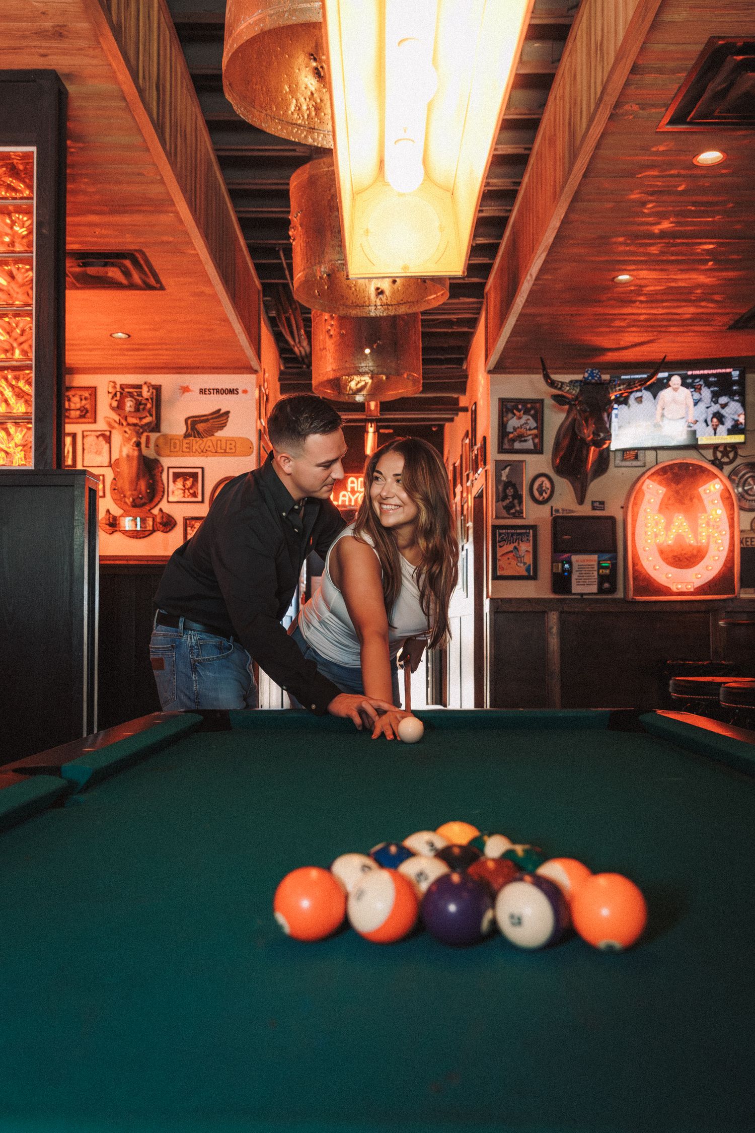 Players strategically line up their shots at a dimly lit pool table with orange pool balls and warm ambient lighting.