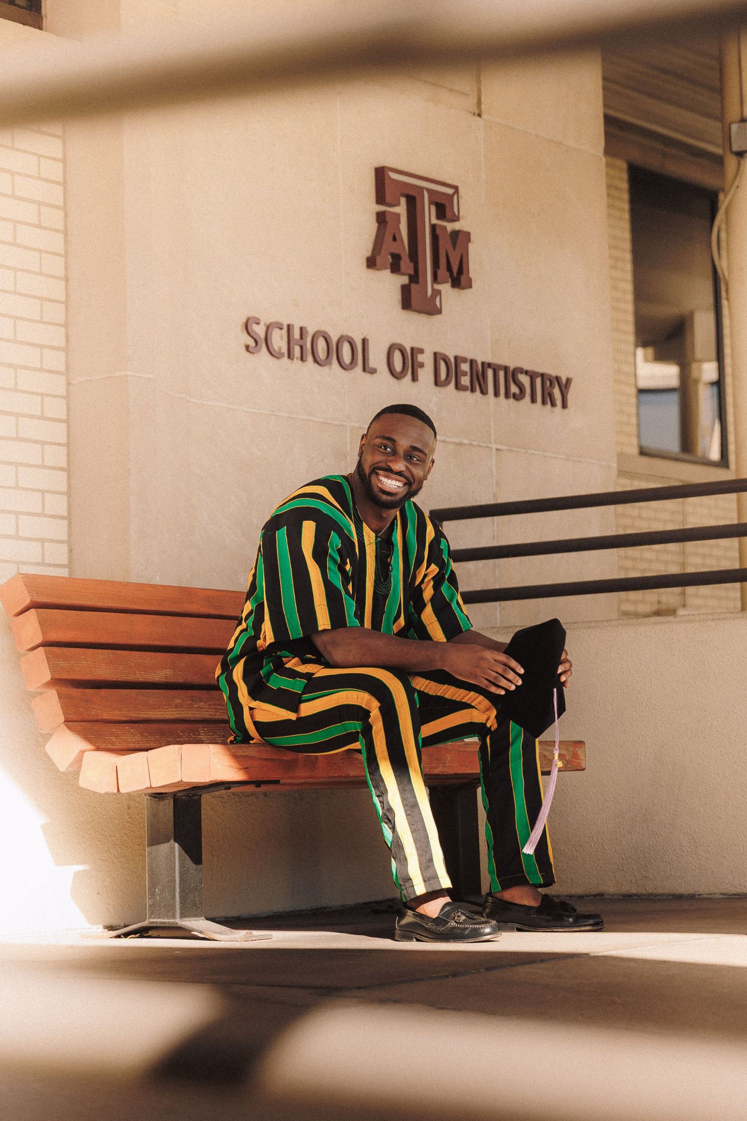 A student in green and black striped attire sits smiling on a bench outside Texas A&M School of Dentistry.