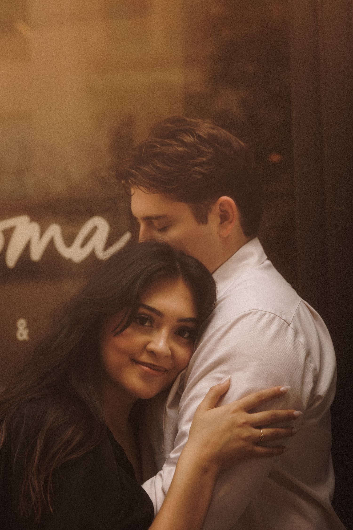 A woman smiles softly as her partner embraces her tenderly in a warm golden-lit romantic moment.