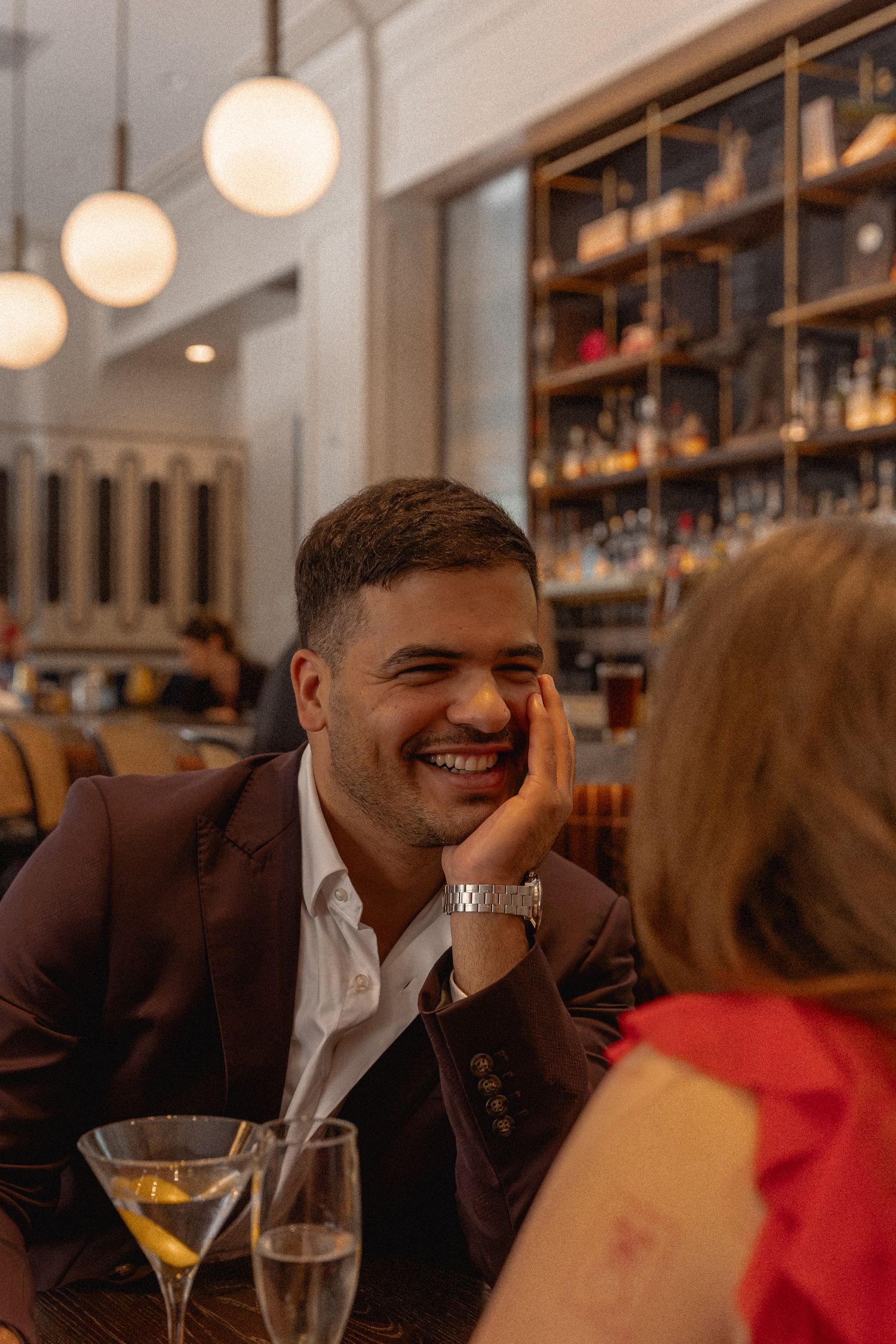 A smiling man in a burgundy blazer enjoys a cocktail at an upscale bar restaurant on a date.