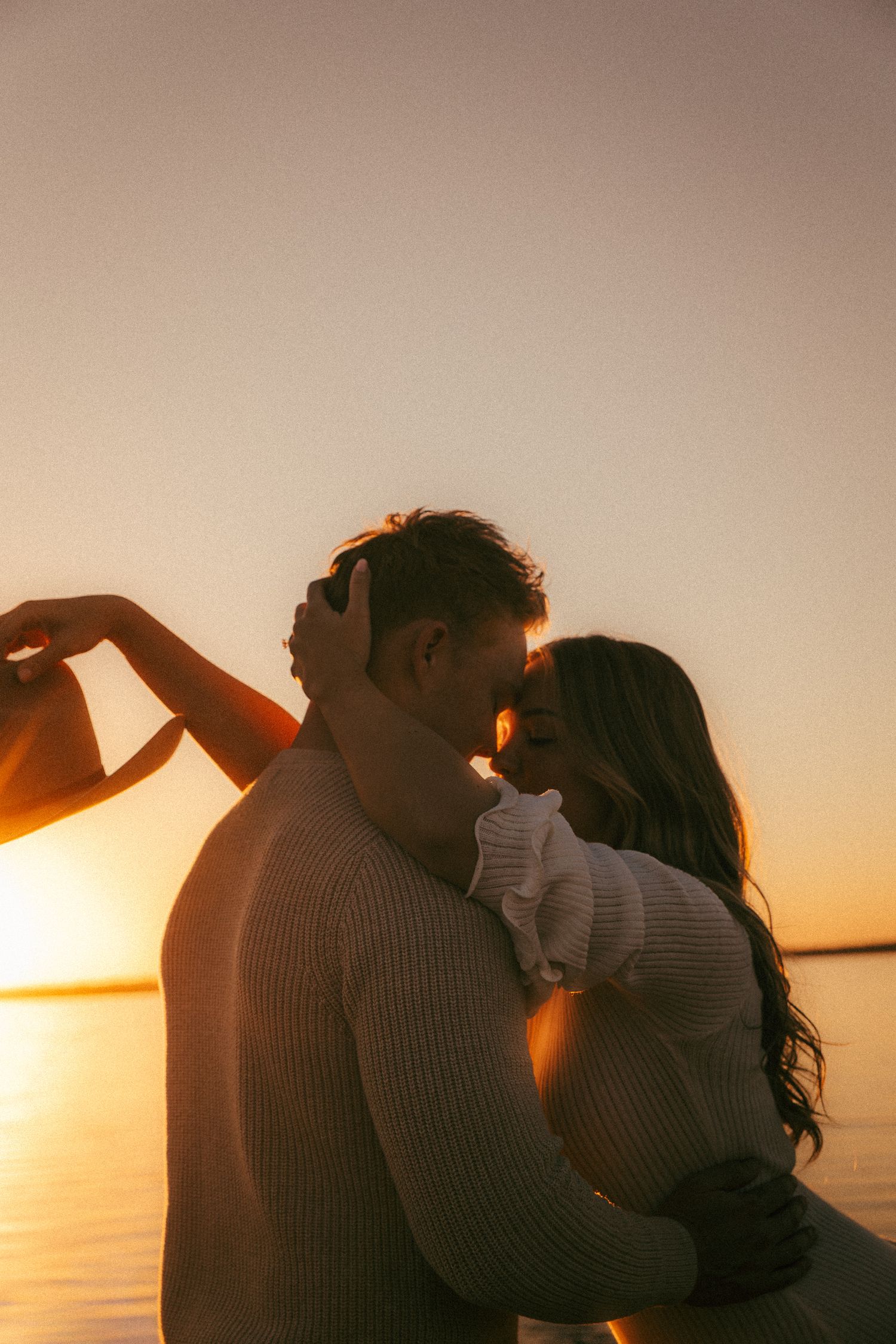 A couple embraces on a beach during golden hour sunset with warm orange light illuminating the romantic scene.