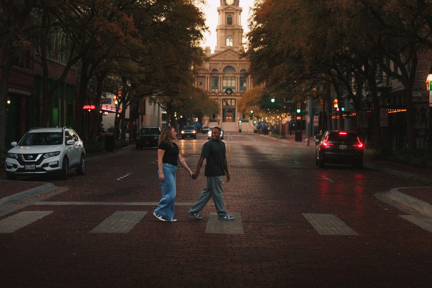 People walking on a city street at dusk with warm lighting and parked cars along the sidewalk.