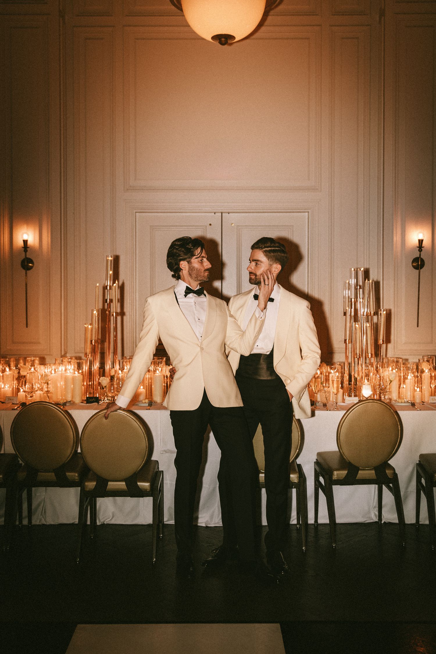 Two grooms in white tuxedo jackets share a romantic moment at their elegant candlelit wedding reception.