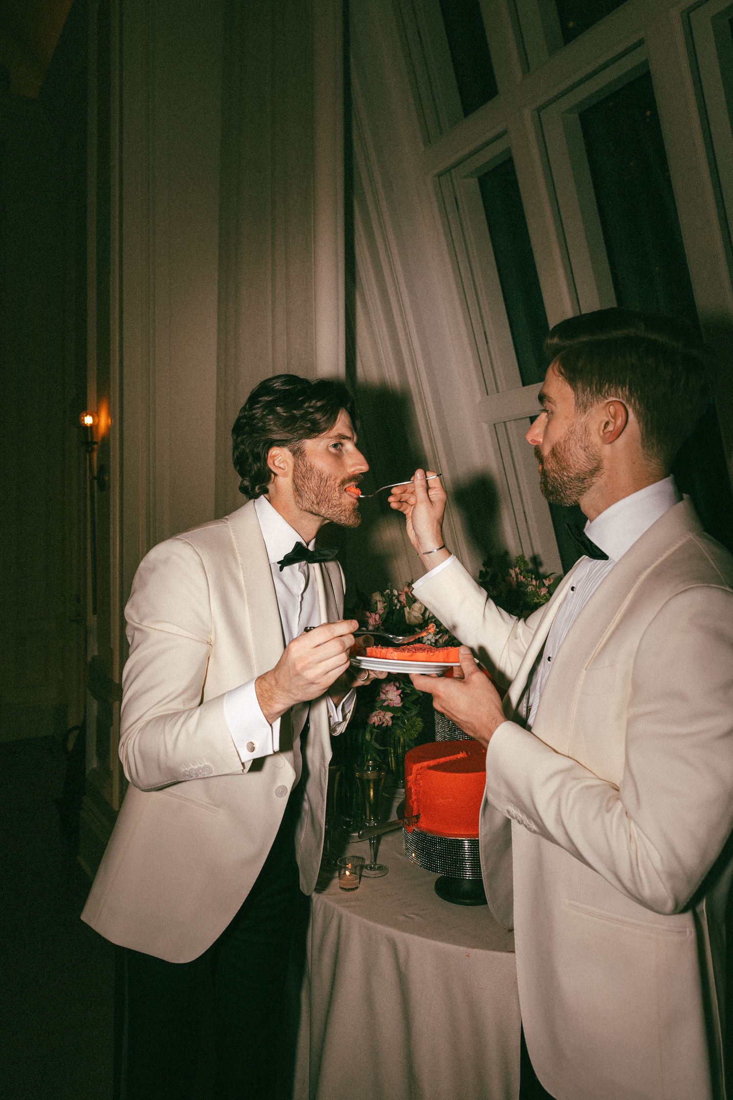 Two grooms in white tuxedos share a playful cake-tasting moment at their wedding reception.