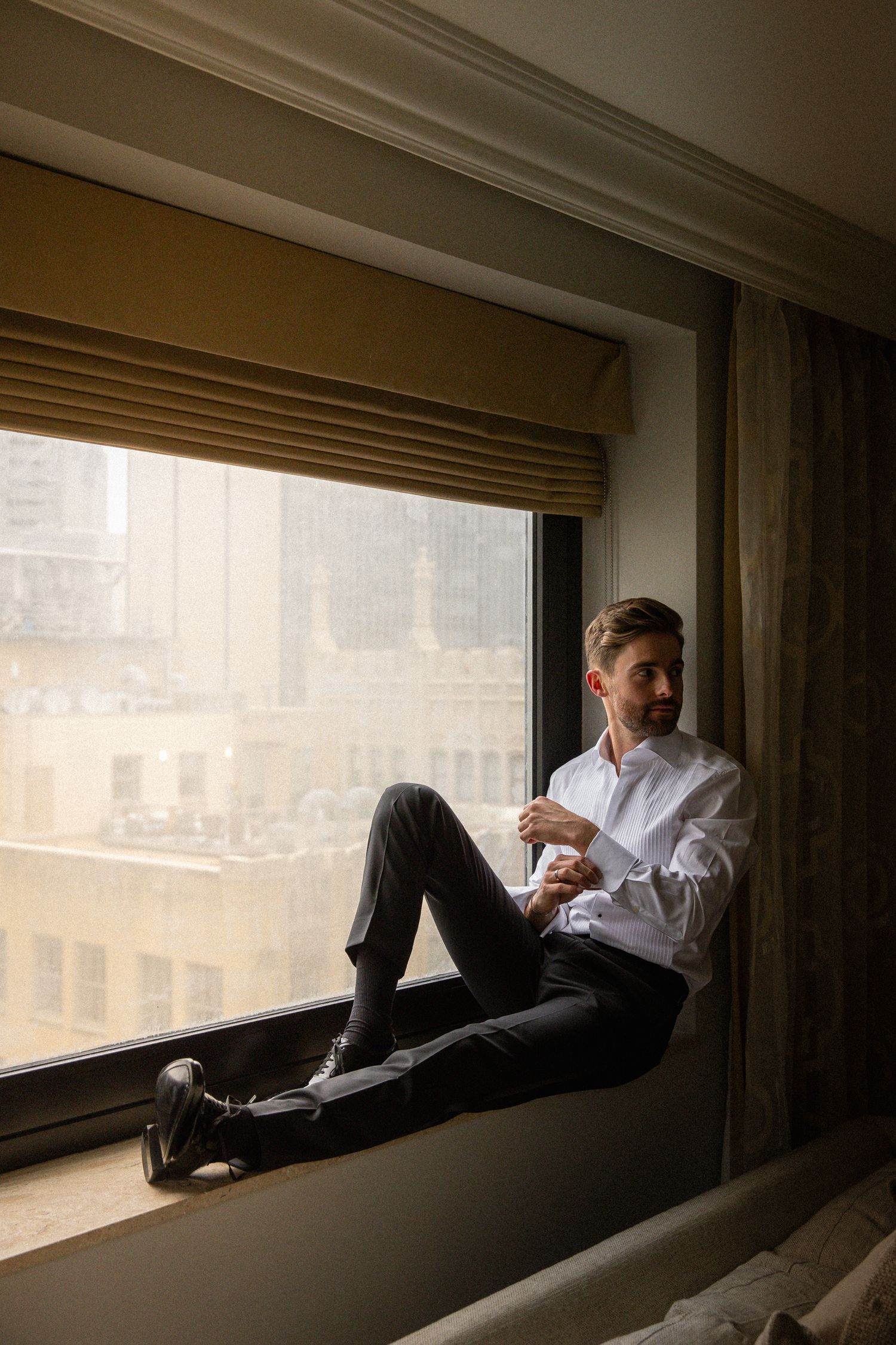 Groom in white dress shirt and black trousers sitting on a hotel window ledge overlooking a city skyline.