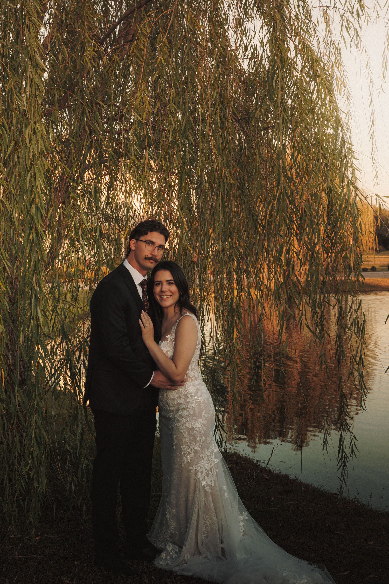 Bride and groom embracing under weeping willow tree by reflective lake at golden hour sunset.