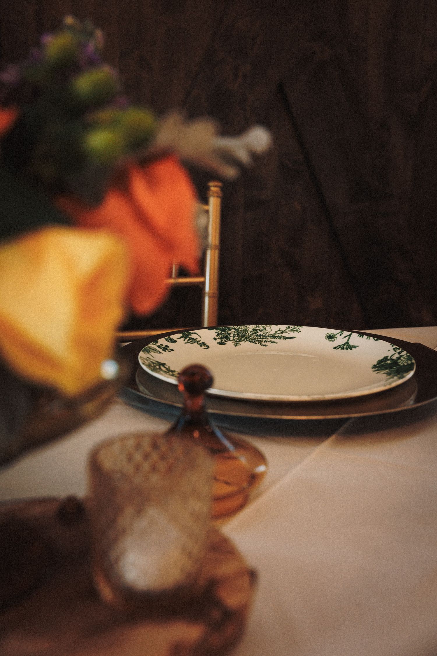 Elegant table setting with green botanical plate, amber glass, gold cutlery, and colorful floral arrangement on white linen.