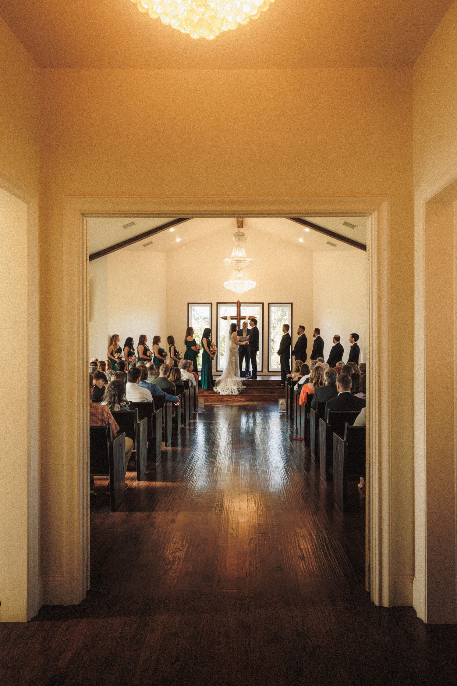 Wide shot of wedding ceremony inside white chapel with guests seated in wooden pews.