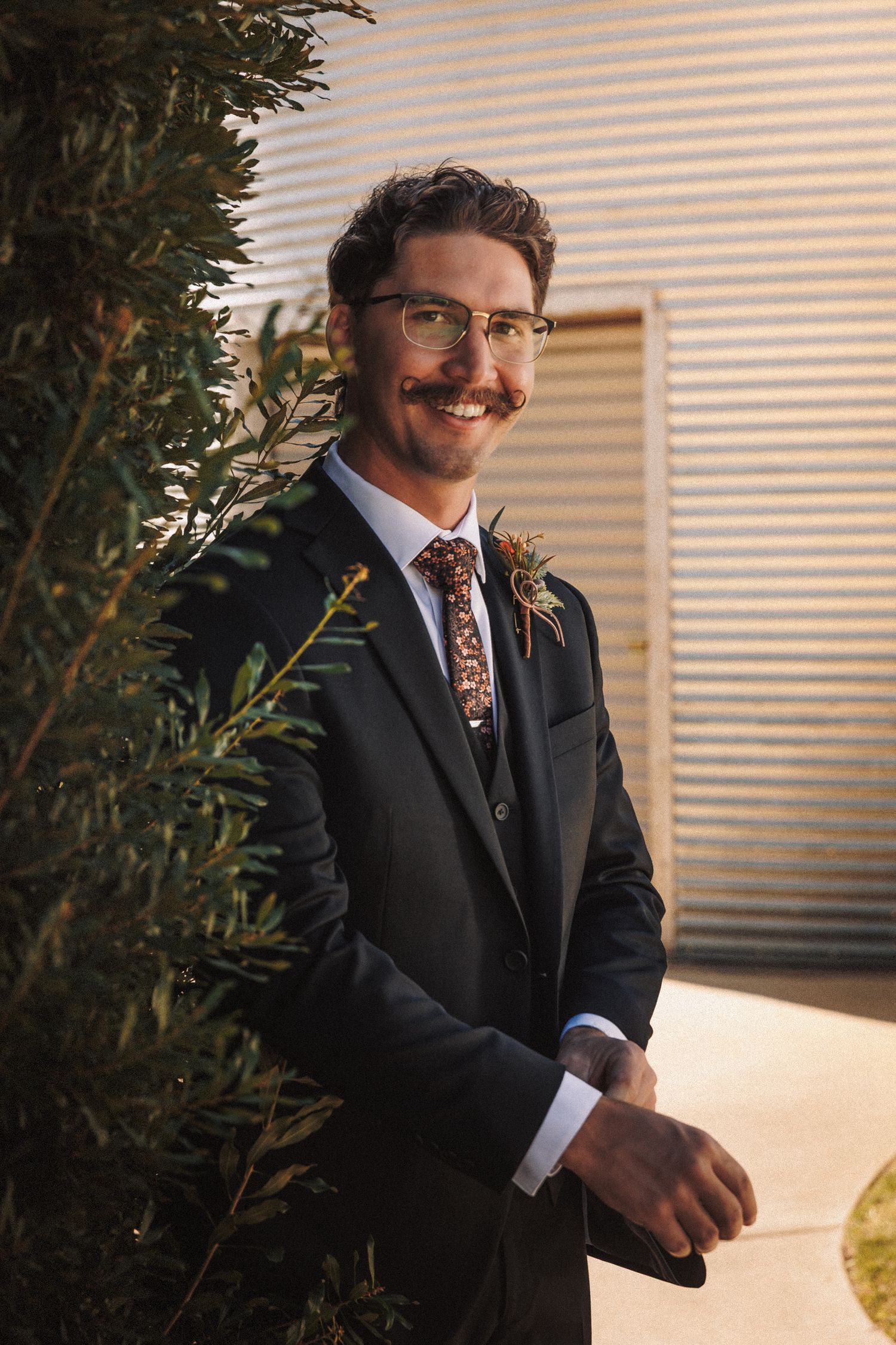 Smiling groom in black three-piece suit with floral tie and boutonniere stands beside green shrubs outdoors.