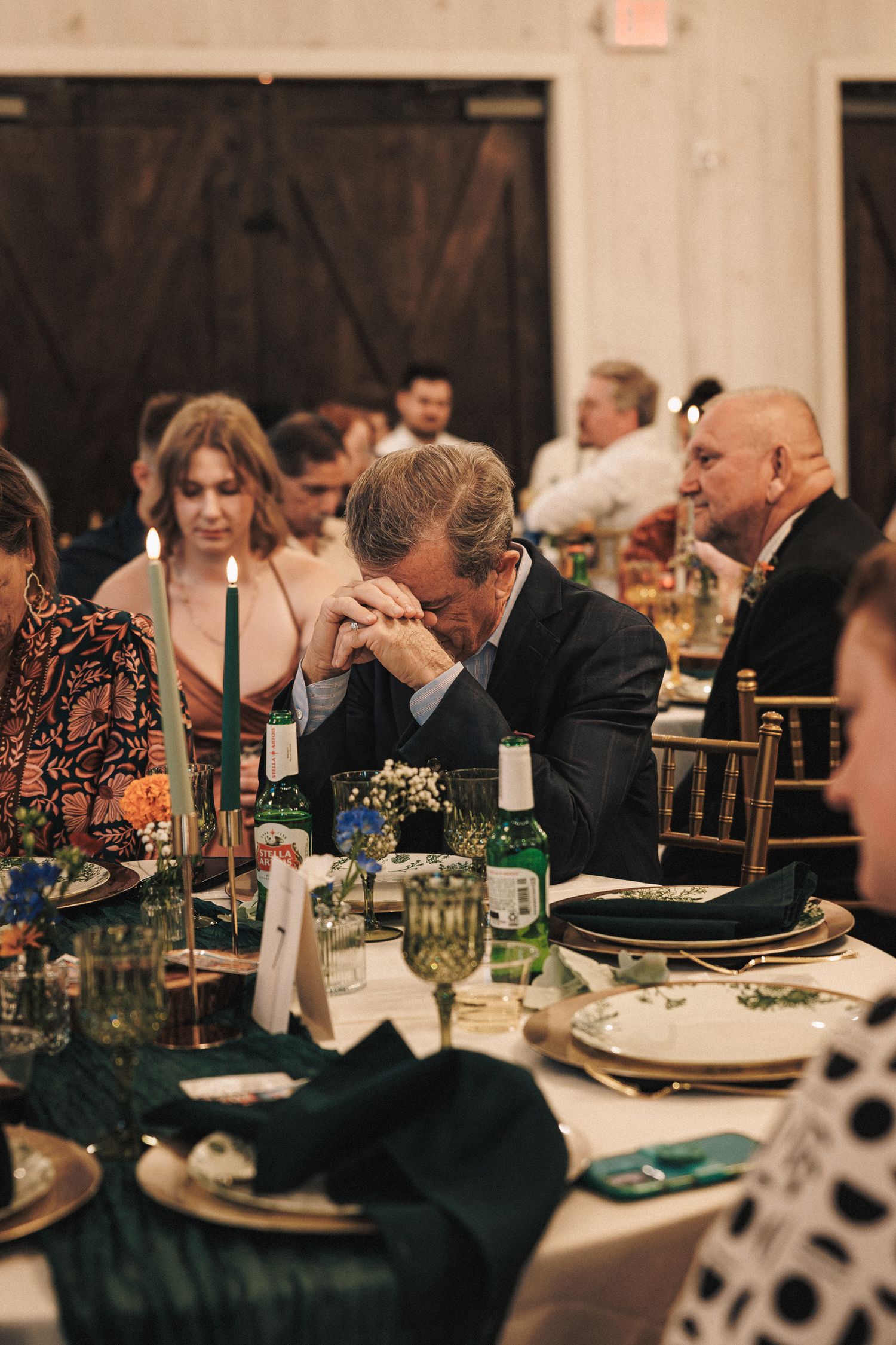 Wedding guests bowing heads in prayer at elegantly decorated reception table with green napkins.