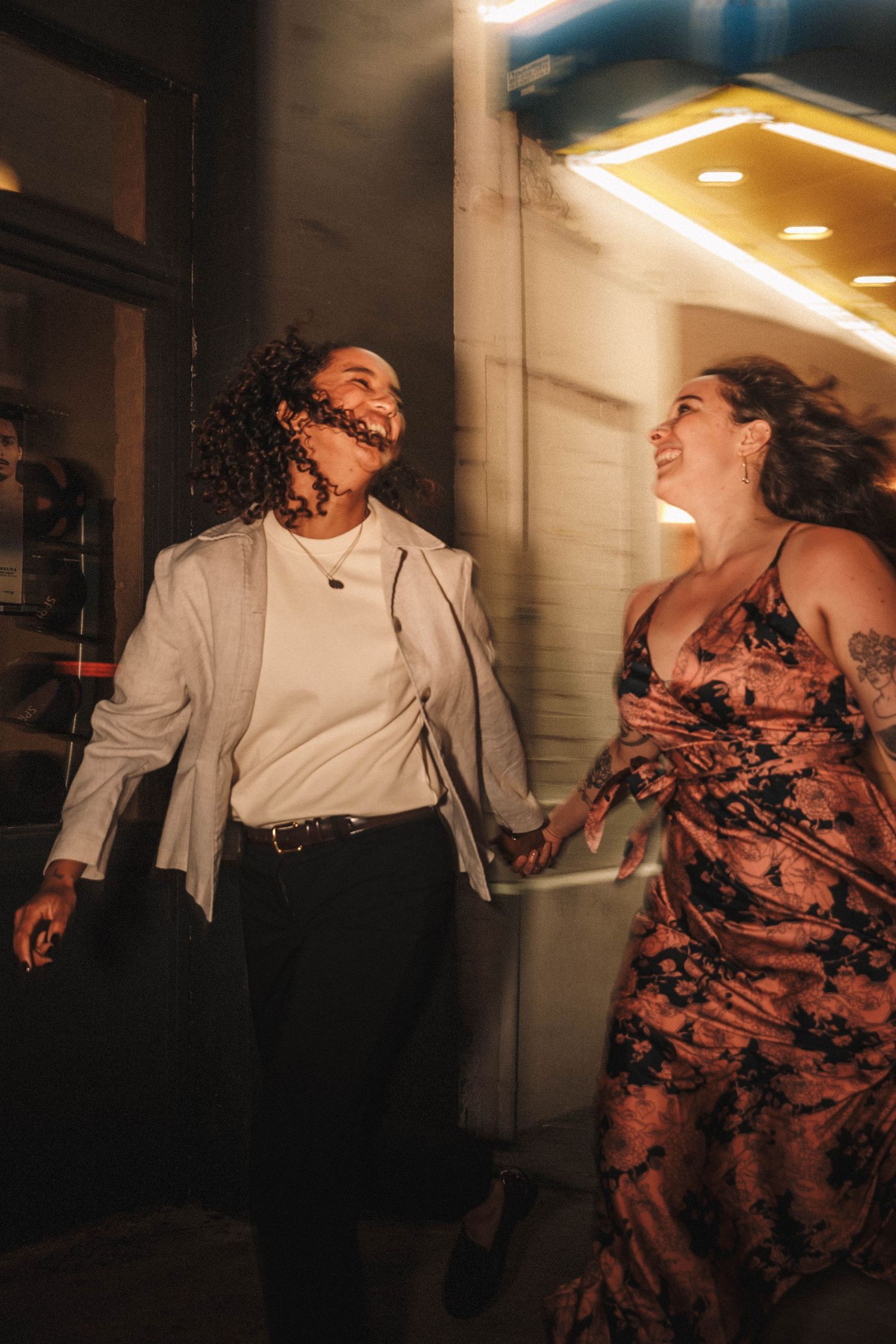 Happy newly engaged couple holds hands laughing while walking outside a theater at night.