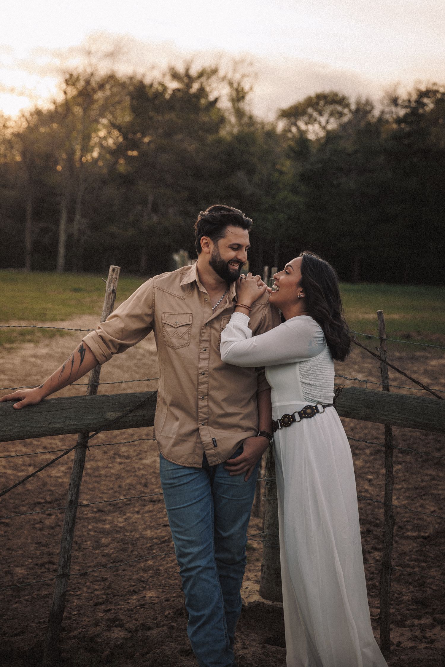 Happy couple laughing together by a wooden fence at sunset on a ranch.