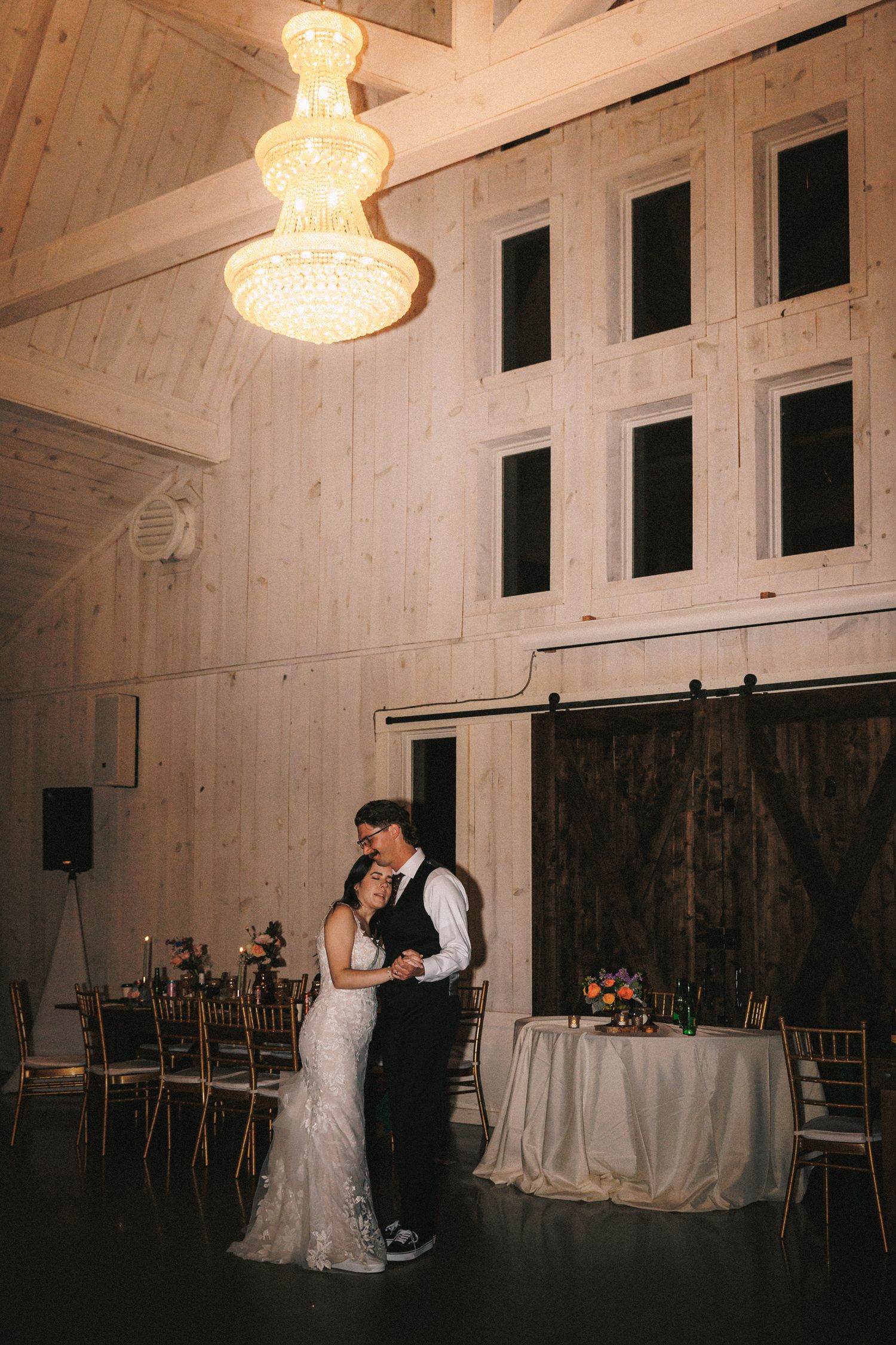 Bride and groom share an intimate first dance beneath a glowing crystal chandelier in a rustic barn wedding venue.