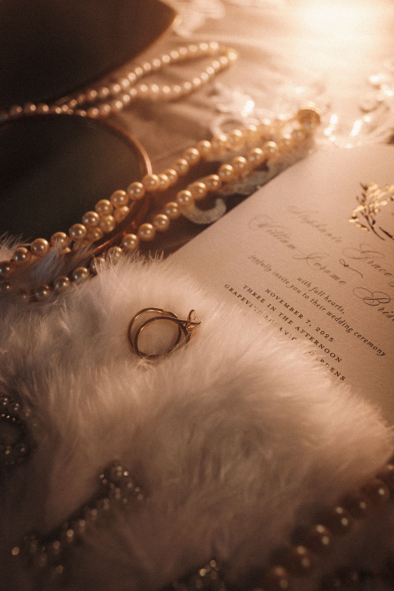 Elegant wedding flat lay with pearl necklaces, gold rings on fur, and a calligraphy invitation in warm golden light.