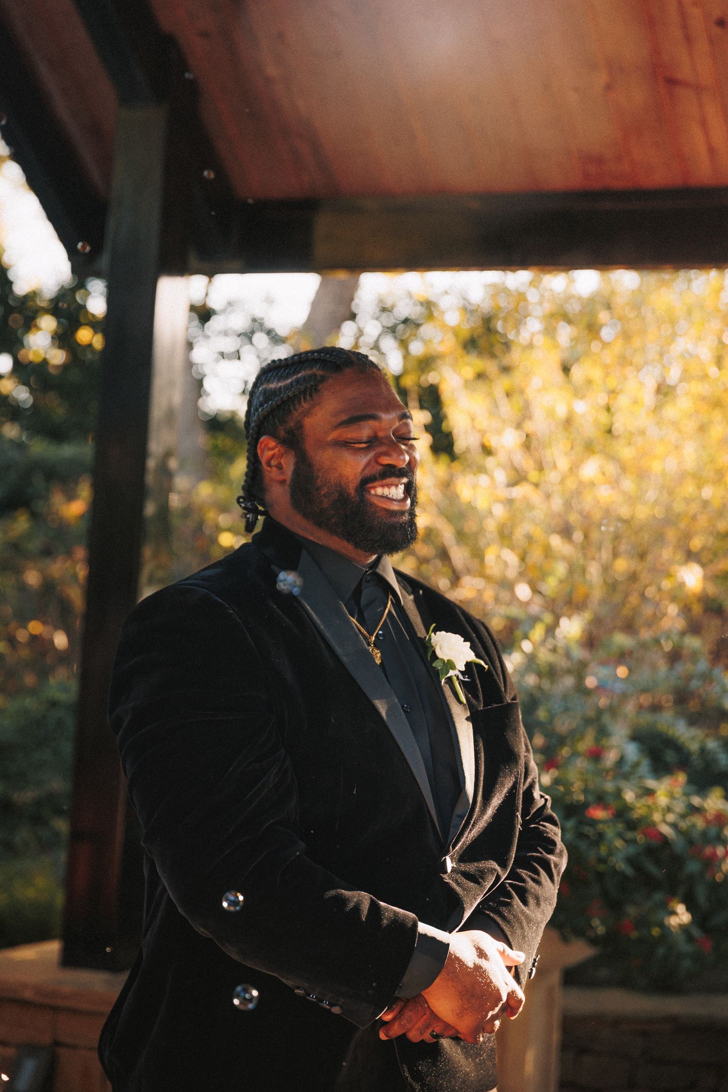 Groom in black velvet tuxedo smiles joyfully under a wooden pavilion surrounded by colorful autumn foliage.