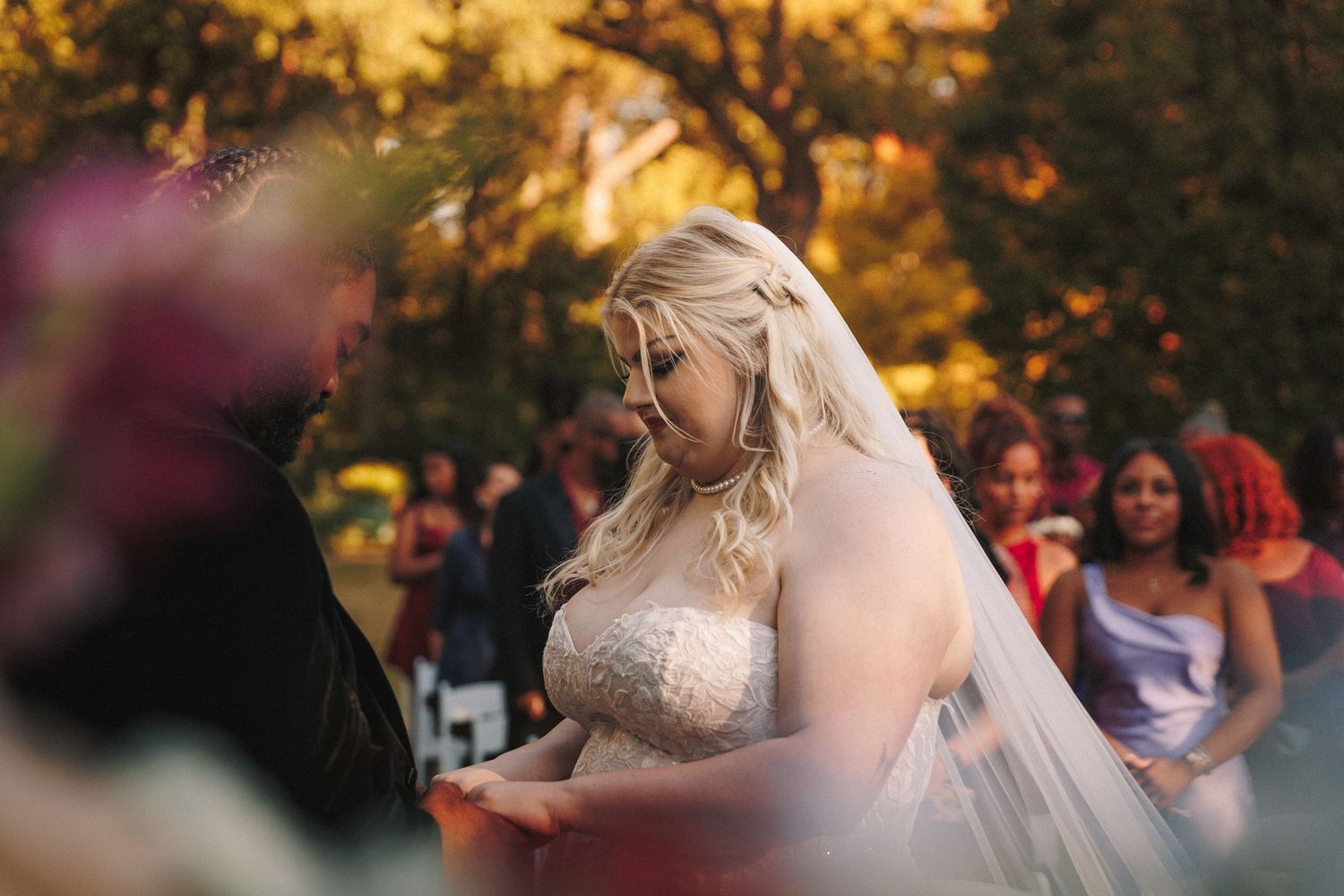 Blonde bride in lace gown and veil exchanging vows at an outdoor autumn wedding ceremony with guests.