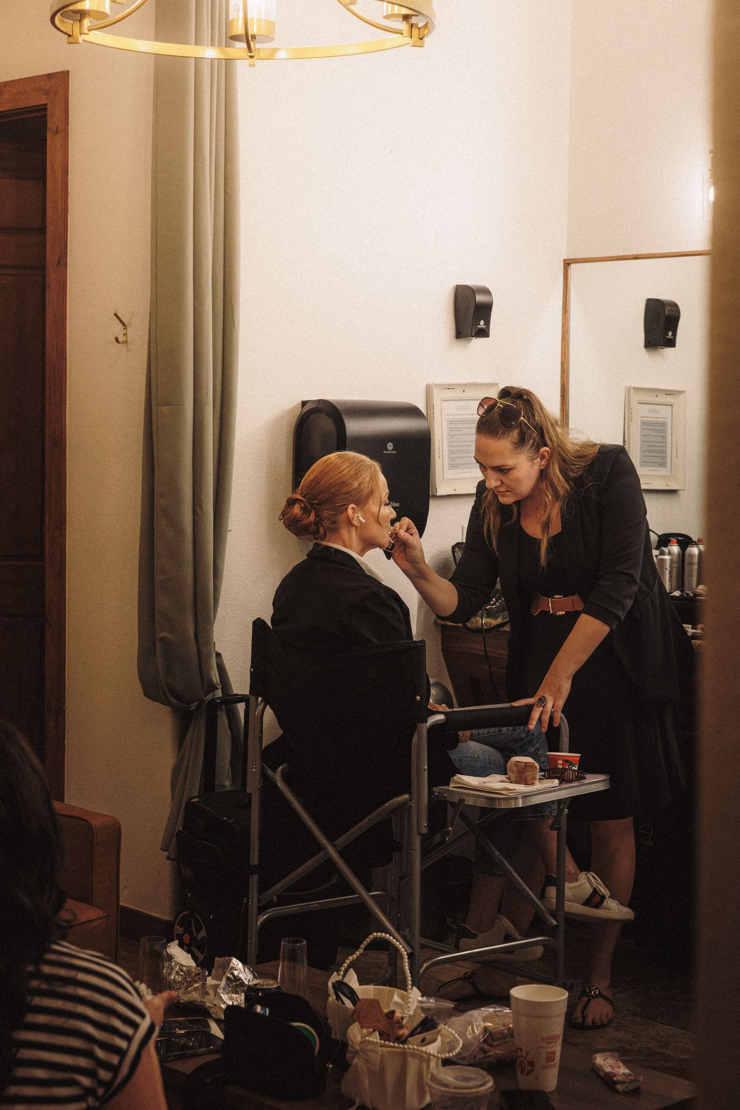 Makeup artist applying cosmetics to a seated woman in a warmly lit bridal preparation room.