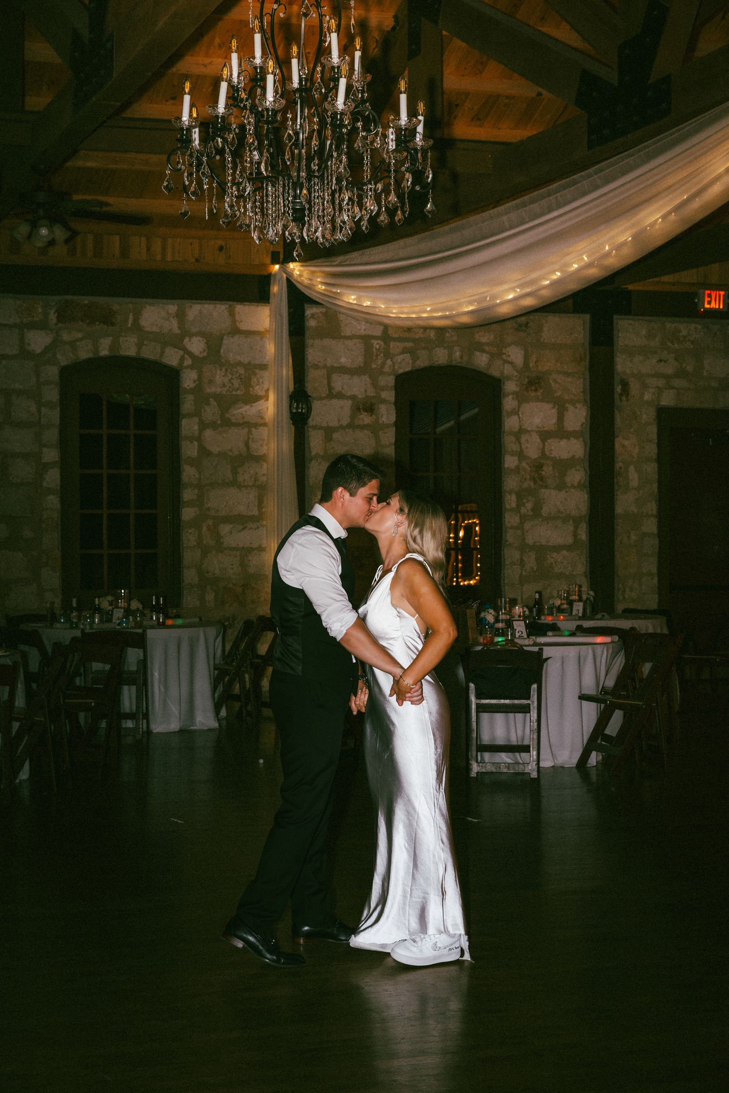 Bride and groom share a kiss during last dance under elegant chandelier in stone wedding venue.
