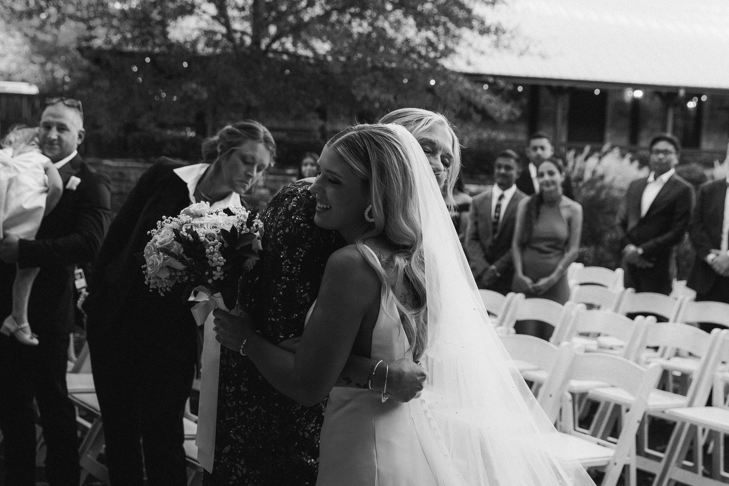 Emotional black and white photo of bride in veil receiving hug during wedding ceremony procession.