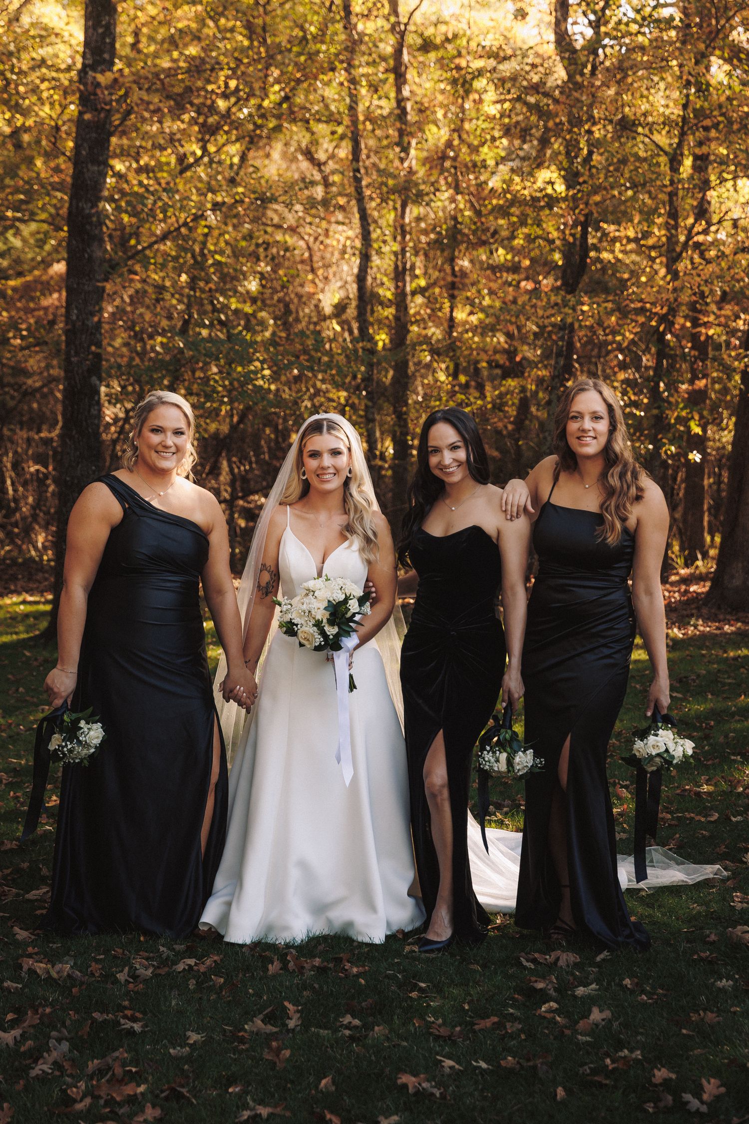 Bride in white gown with three bridesmaids in black dresses holding white bouquets in autumn forest.