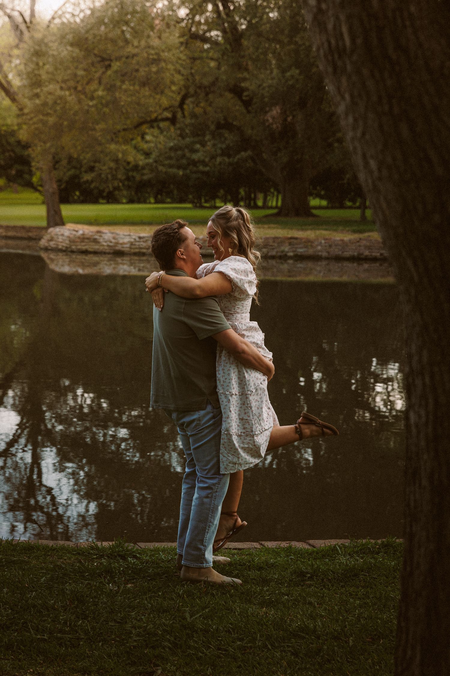 Man joyfully lifting woman in white dress by a serene reflective pond surrounded by lush green trees.