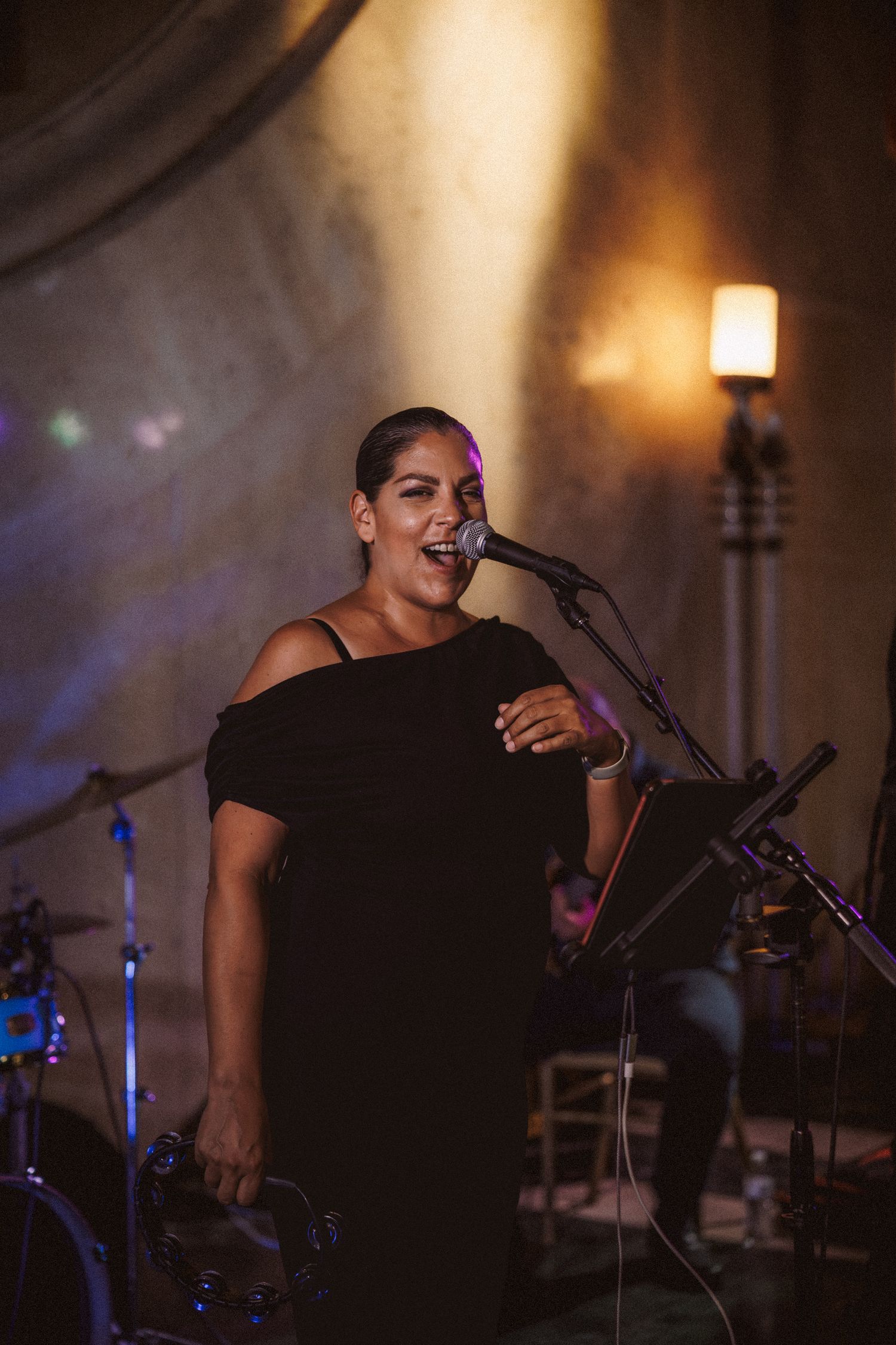 Female vocalist in black dress performing at microphone stand under warm stage lighting.