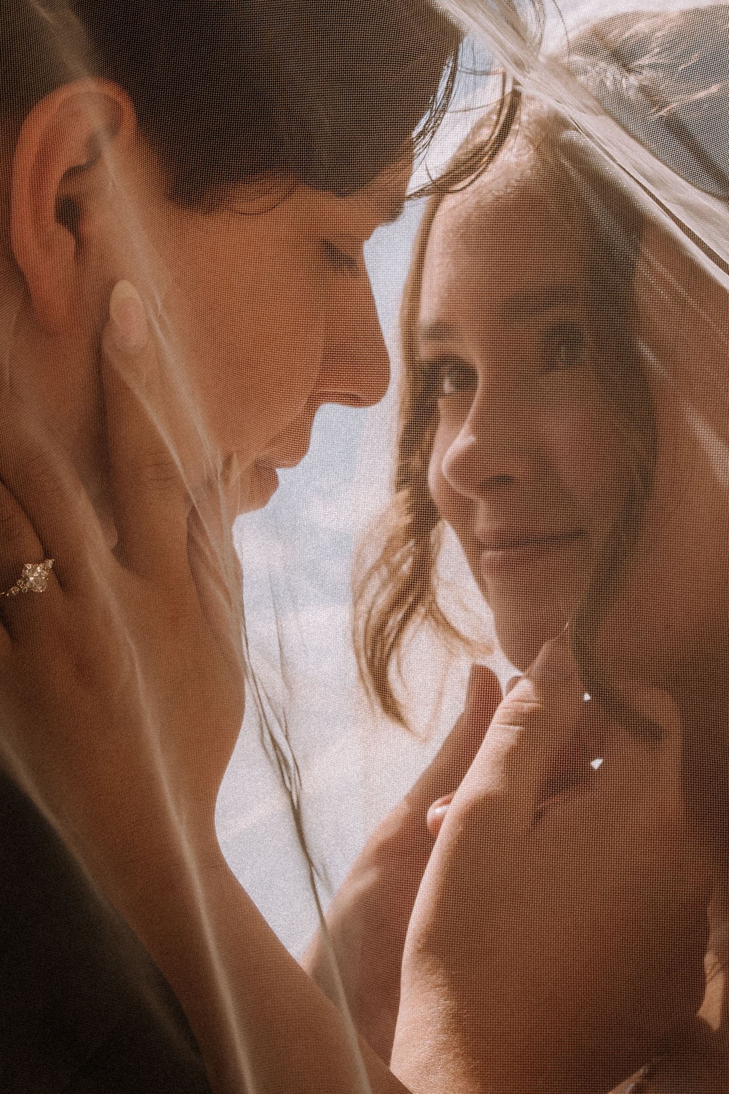 Two brides share an intimate moment under a wedding veil, one touching the others face tenderly.
