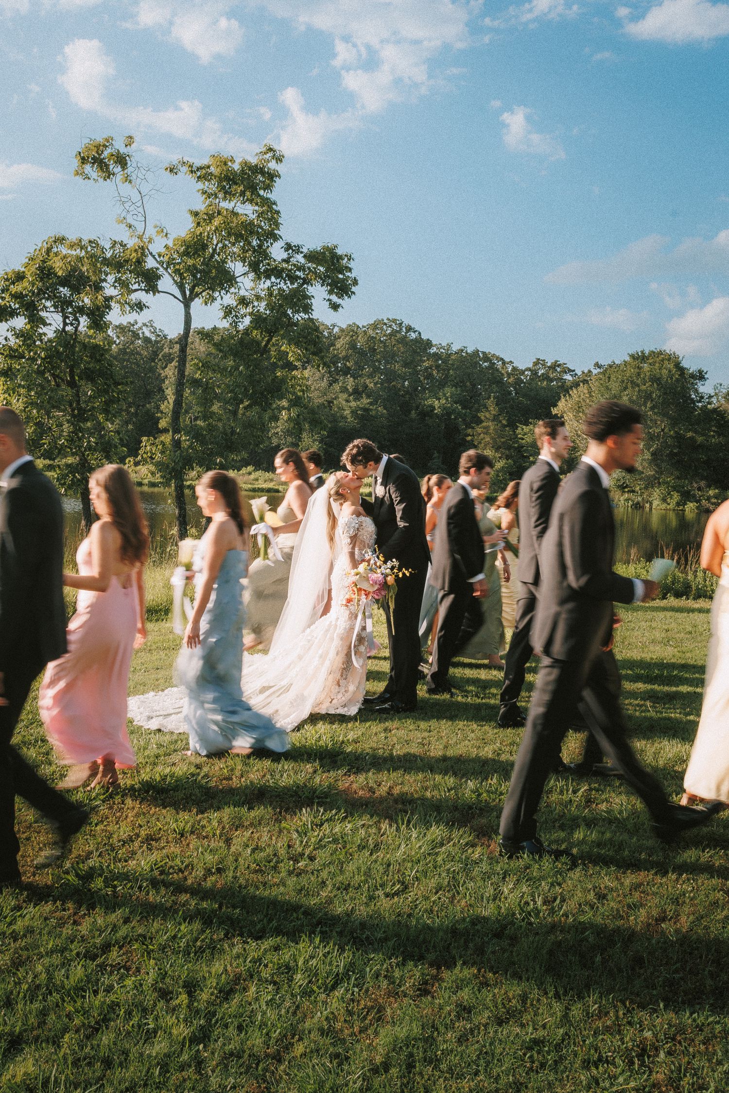 A wedding party walking through a grassy field under a blue summer sky.